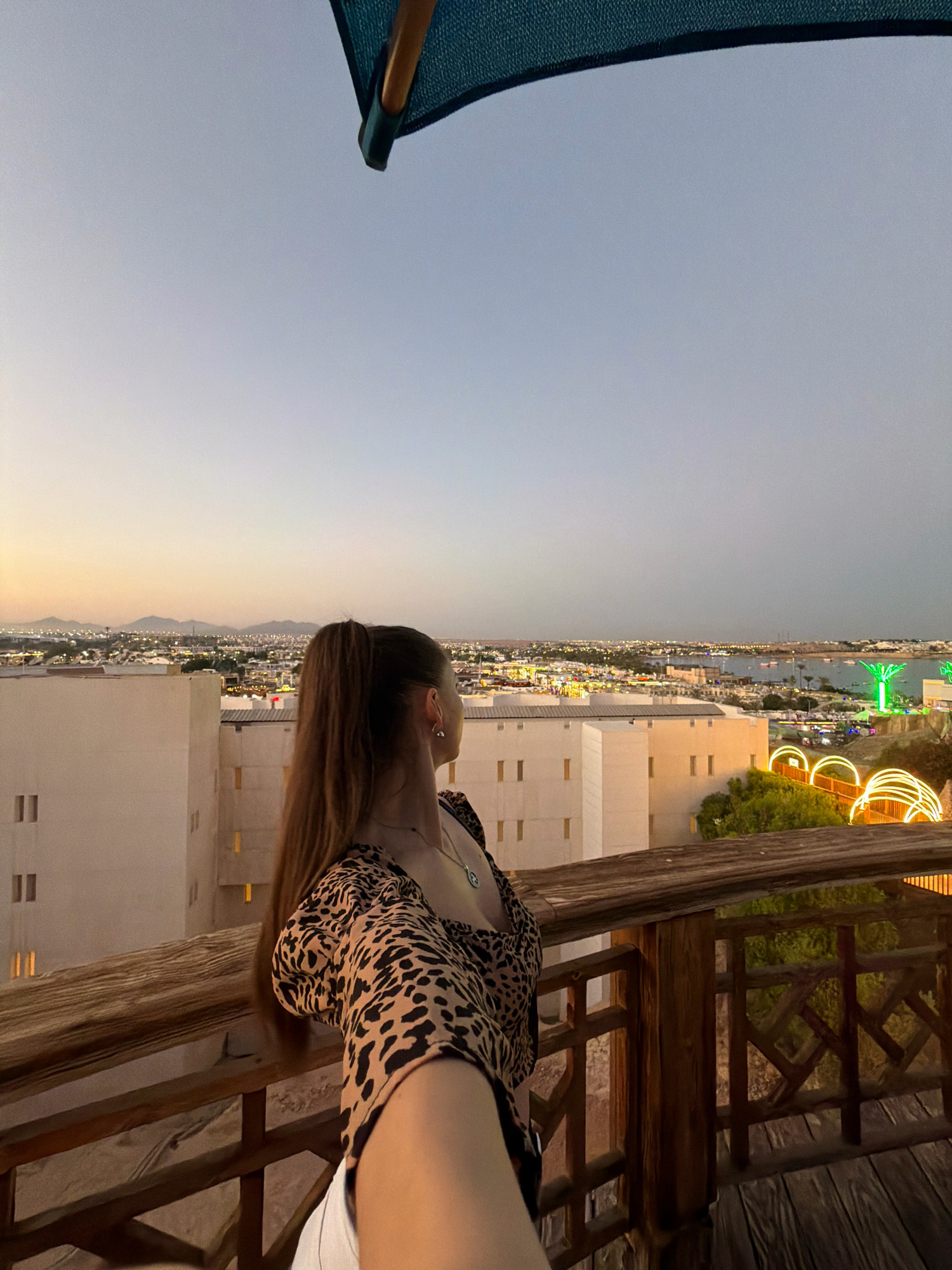 The image captures a woman standing on a wooden balcony, looking out at a panoramic view of Sharm El Sheikh, Egypt, at dusk. The woman, facing away from the camera, has her hair in a high ponytail and is wearing a leopard-print top and white shorts. The city lights twinkle below, illuminating buildings and the bay. In the foreground, the wooden railing of the balcony is visible, along with parts of the woman's arm and shoulder. The sky is a gradient of soft blues and oranges, suggesting the transition from day to night. The overall scene conveys a sense of tranquility and the beauty of the coastal city.