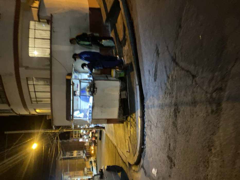 The image depicts a nighttime street scene in La Paz, Bolivia. A white, mobile food cart with an illuminated interior is positioned on a concrete sidewalk at a street corner, adjacent to a two-story building with a curved, light-colored facade and dark-trimmed windows.
Four individuals are present: one person, wearing a blue top, is inside the food cart, appearing to operate it. Three customers stand to the right of the cart. The first customer, wearing a dark blue jacket and pants, is closest to the cart, facing the vendor. The second customer, wearing a dark blue jacket and a dark baseball cap, stands behind the first. The third customer, wearing a green jacket, stands furthest right, holding a light-colored bag or object.
The street, constructed of asphalt, shows cracks and wear. A concrete curb separates the sidewalk from the road. A yellow streetlight on a utility pole with numerous overhead wires illuminates the left side of the scene. Several vehicles are parked further down the street in the background. A yellow bucket is visible next to the food cart. A piece of white debris rests on the street in the foreground.