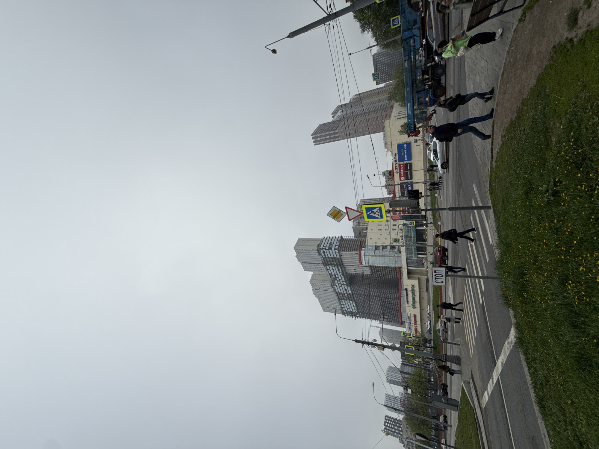 The image shows a street scene in Moscow, Russia. Several high-rise buildings are visible in the background. Pedestrians are crossing the street at a crosswalk. The street has a "STOP" sign in Russian ("СТОП"). Traffic lights and street lights are also present. A construction crane and several cars are parked along the street. The sky is overcast.