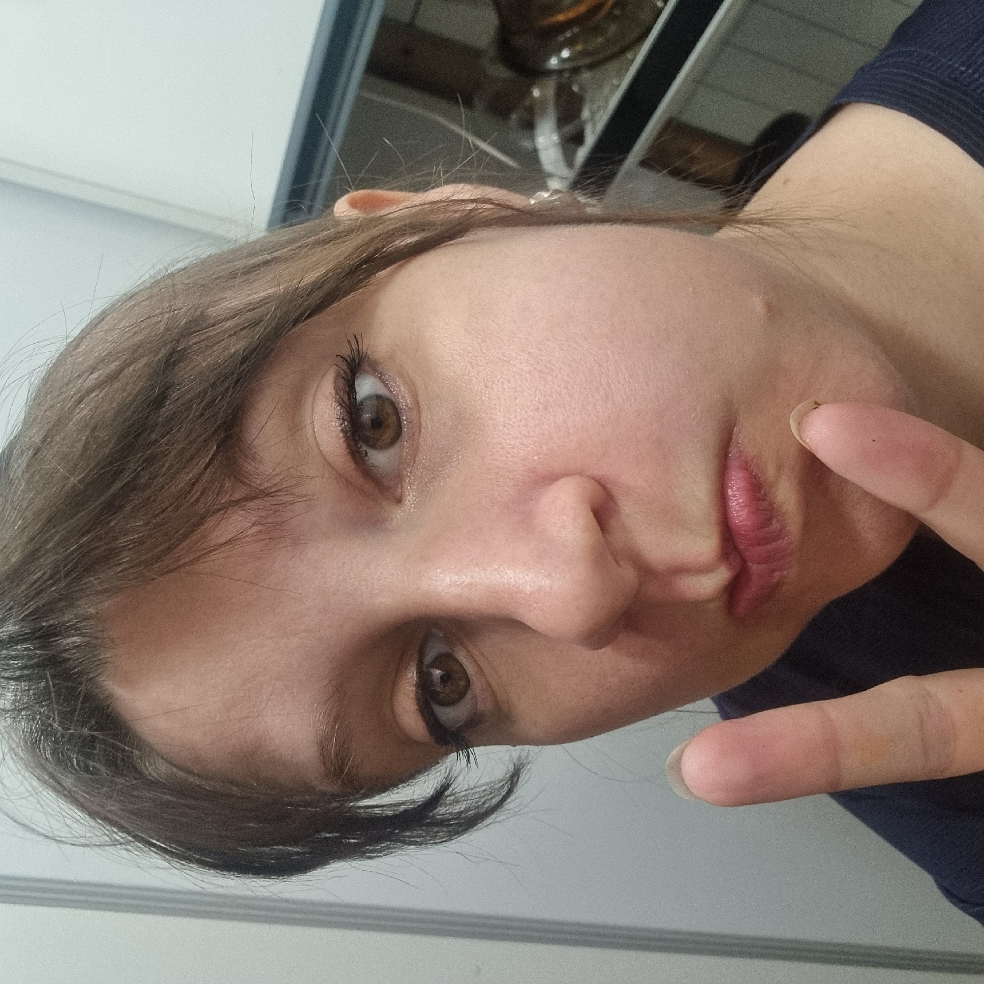 The image shows a close-up, selfie-style photograph of a person's face. The person has light skin, brown eyes, and brown hair. Their lips are slightly pursed, and they are holding up two fingers in a peace sign near their mouth. The background appears to be indoors, with a white wall or cabinet to the left and a darker, reflective surface with some objects to the right, possibly a kitchen or appliance. No specific location in Dunnae-myeon, South Korea, can be identified from the image.