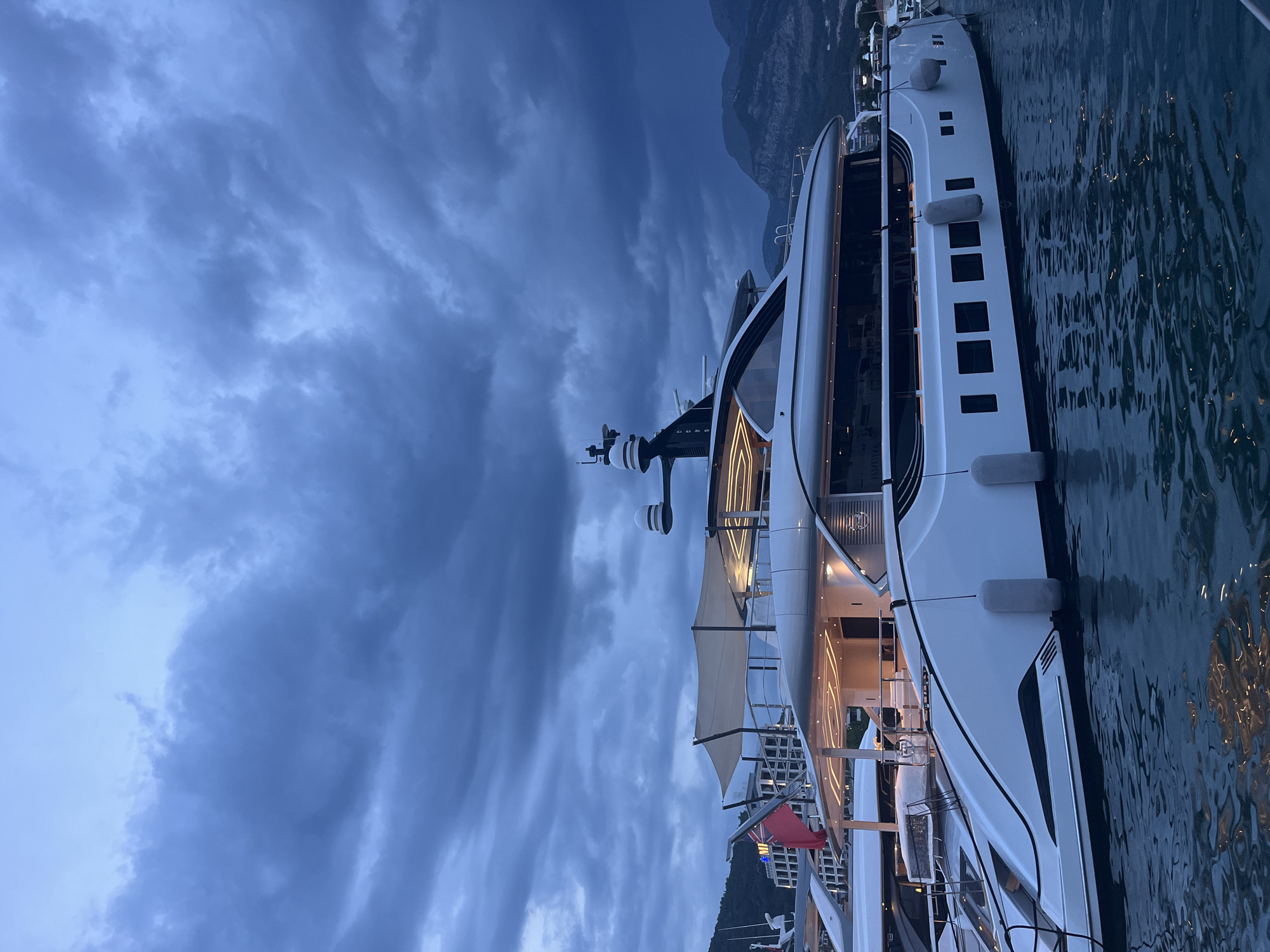 A large, modern white yacht is docked in the waters of Budva, Montenegro. The yacht features multiple levels, sleek lines, and numerous windows. A red flag is visible on one of the upper decks. The sky above is dark and overcast with heavy clouds, suggesting impending rain or a storm. In the background, mountains rise behind buildings lining the shore. The water is calm, reflecting the yacht and the moody sky. There are no people visible in the image.