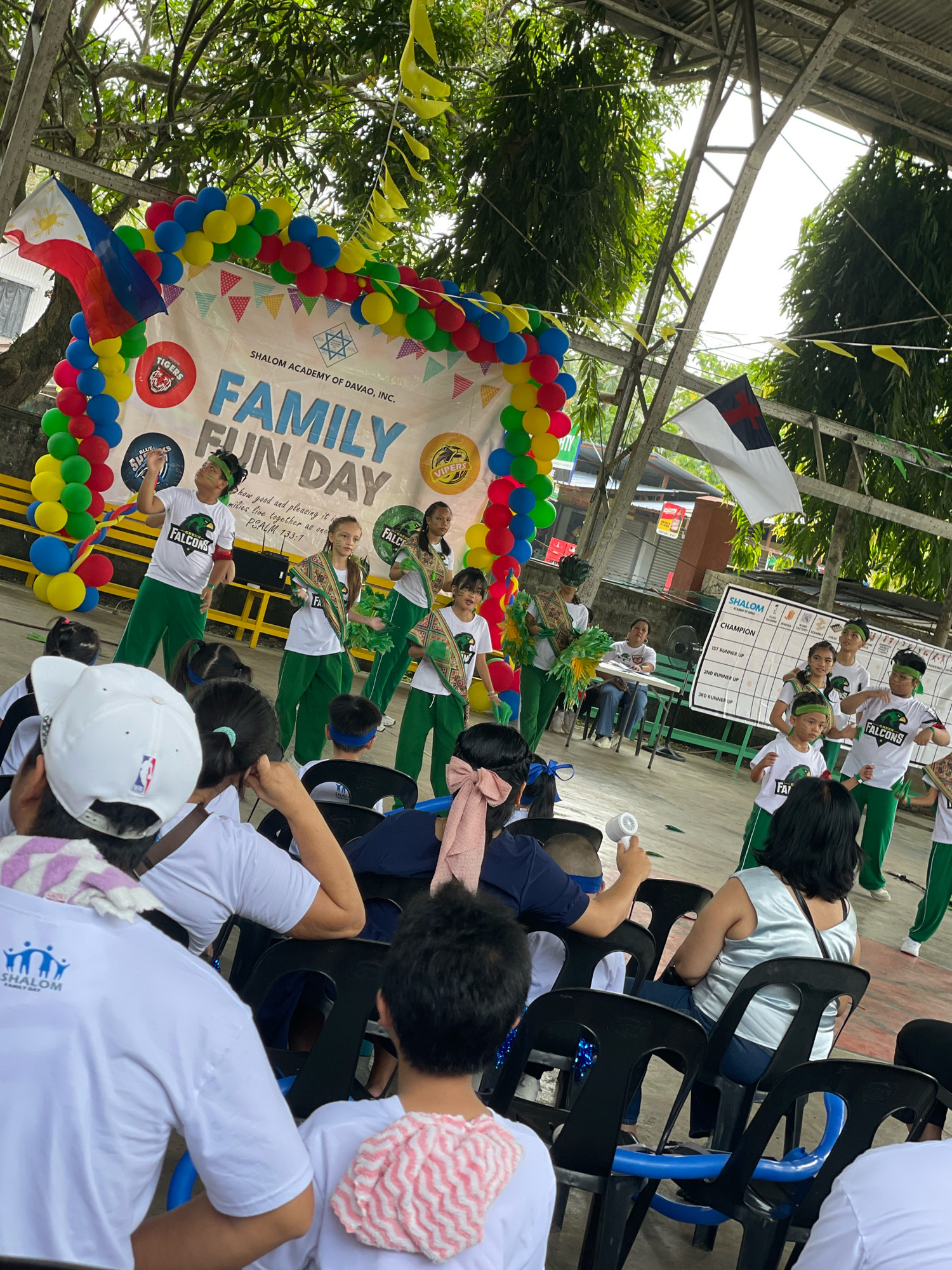 In Davao City, Philippines, an outdoor event, possibly at a school, is underway. A banner displaying "SHALOM ACADEMY OF DAVAO, INC. FAMILY FUN DAY" is prominently featured, adorned with colorful balloons. A group of children in matching white shirts and green pants are performing on a stage, some holding green pom-poms and wearing decorative sashes. An audience, including adults and children, are seated in black plastic chairs, observing the performance. Some audience members wear white t-shirts with the "SHALOM FAMILY DAY" logo. Two flags are visible: one is the Philippine flag, and another is a white flag with a red cross. The background shows trees and a covered structure, suggesting an open-air venue or a covered court. The lighting indicates it is daytime.