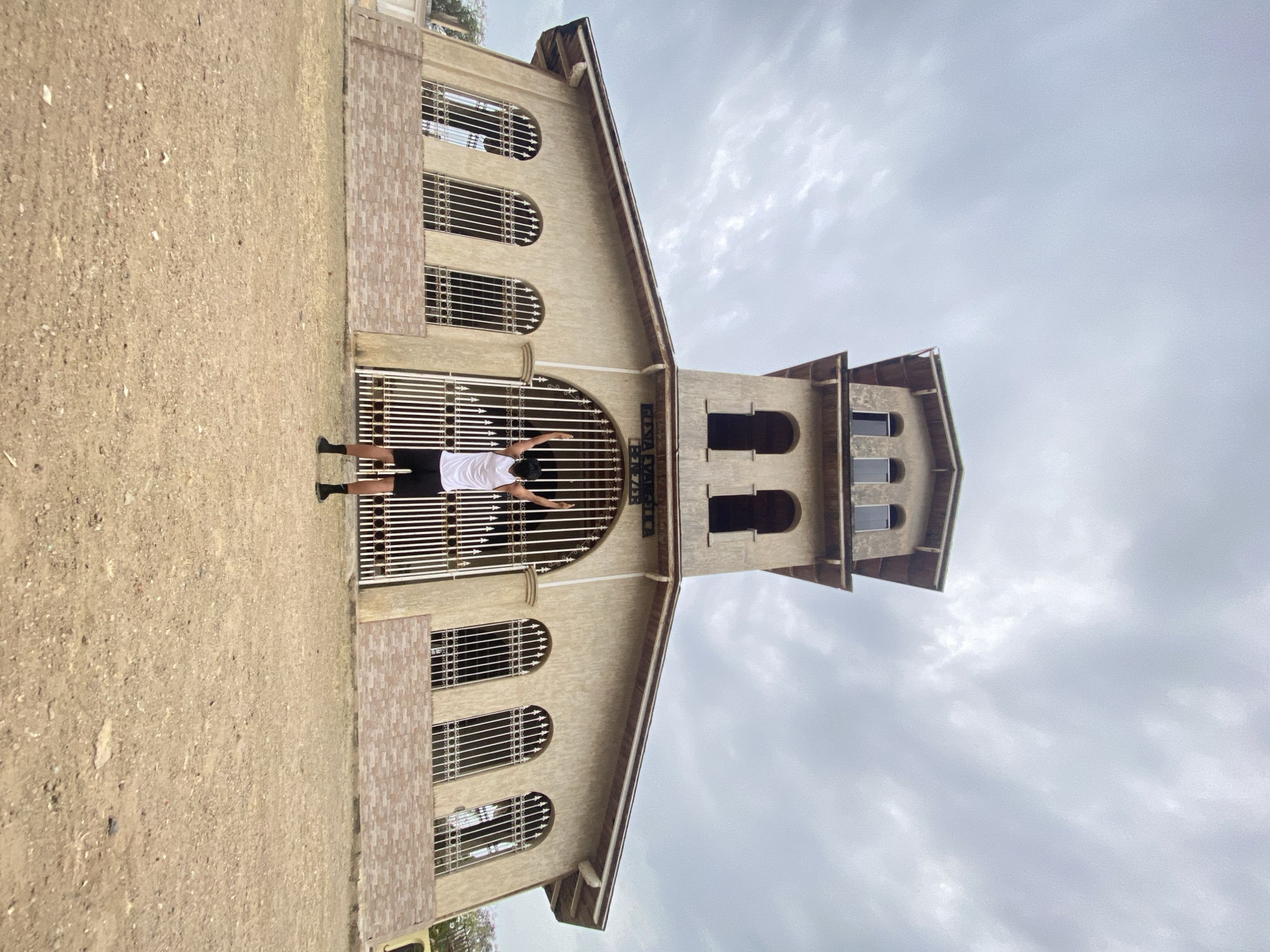 A full-shot photograph captures a light-colored building with a single person standing before its main entrance on an overcast day.
The central subject is a person, likely male, dressed in a white t-shirt, black shorts, and dark shoes or socks. He stands with his back to the viewer, facing the building's large metal gate, with his arms outstretched horizontally, hands resting on the gate's bars. This pose conveys an expressive, possibly welcoming or joyful gesture.
The building itself is a light tan or beige structure, potentially a church or community building, characterized by a large arched main entrance. This entrance is secured by a white, decorative metal gate featuring horizontal bars and scrollwork. Above the arch, a sign displays text. To the left and below the main arch, the facade incorporates three more arched windows, each fitted with matching metal grates. To the right of the main building, a prominent tower rises, featuring several smaller arched openings on its upper sections. The building's rooflines are gabled.
The foreground consists of unpaved, compacted dirt or gravel. The sky is uniformly covered with light grey clouds, indicating overcast weather.
Visible text on the sign above the main entrance gate clearly reads "IGLESIA EVANGELICA NAZARENA". The scene suggests a local community or religious building in Juan Griego, Venezuela.