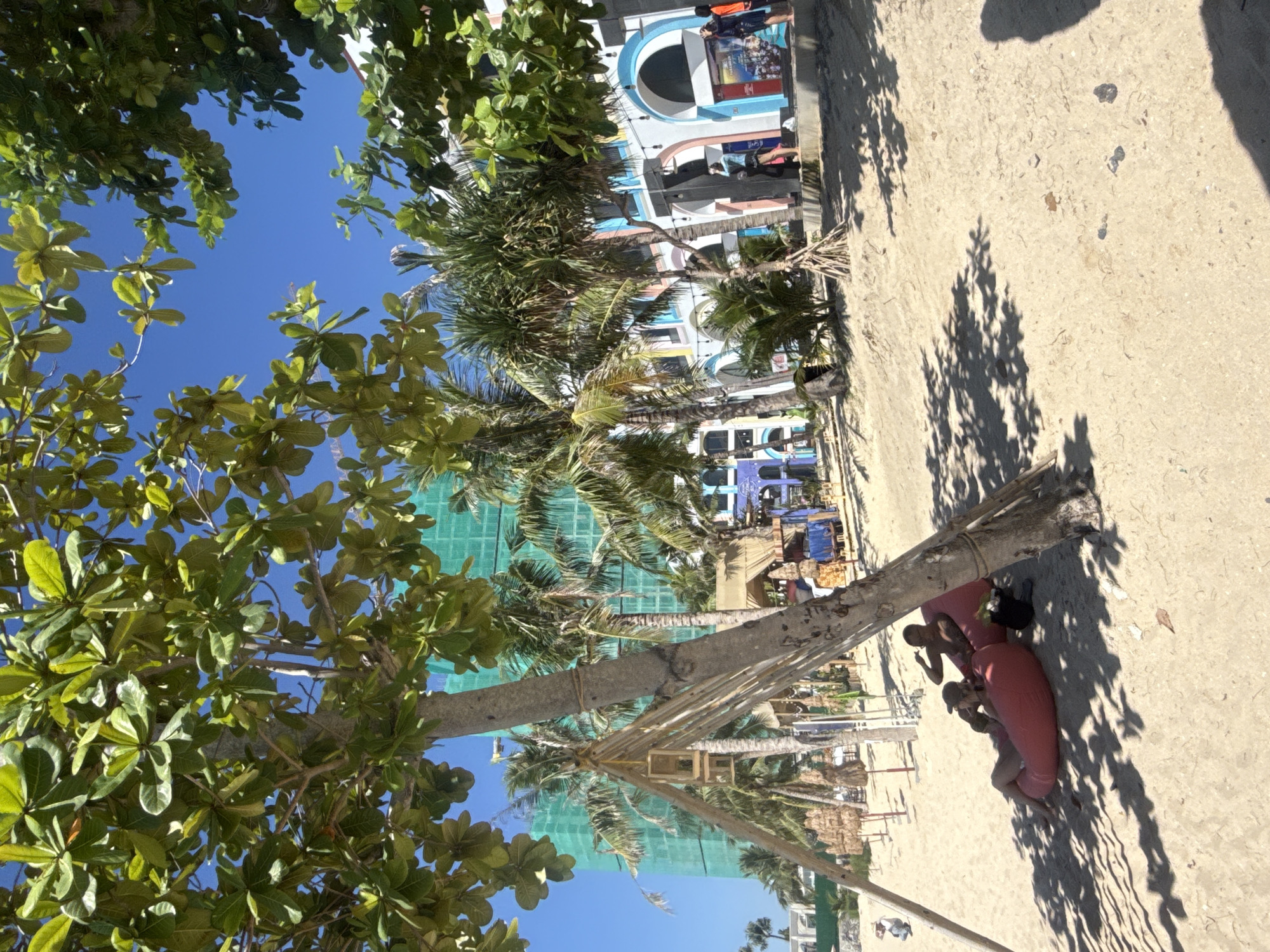 This image captures a scene at a beach in Nha Trang Ward, Vietnam. In the foreground, a large tree with green leaves partially obscures the view of a sandy beach where two people are relaxing on pink beanbag chairs. One person is lying down, holding a drink, while the other sits up, looking towards the right. Behind them, a structure made of bamboo and wood is visible, possibly part of a beach shelter. Further in the background, palm trees and a building under construction, draped in green netting, dominate the landscape. The building features colorful arches and windows, suggesting it's a resort or hotel. Other beach amenities like thatched umbrellas and small stalls are also present. The sky is a clear blue, indicating a sunny day.