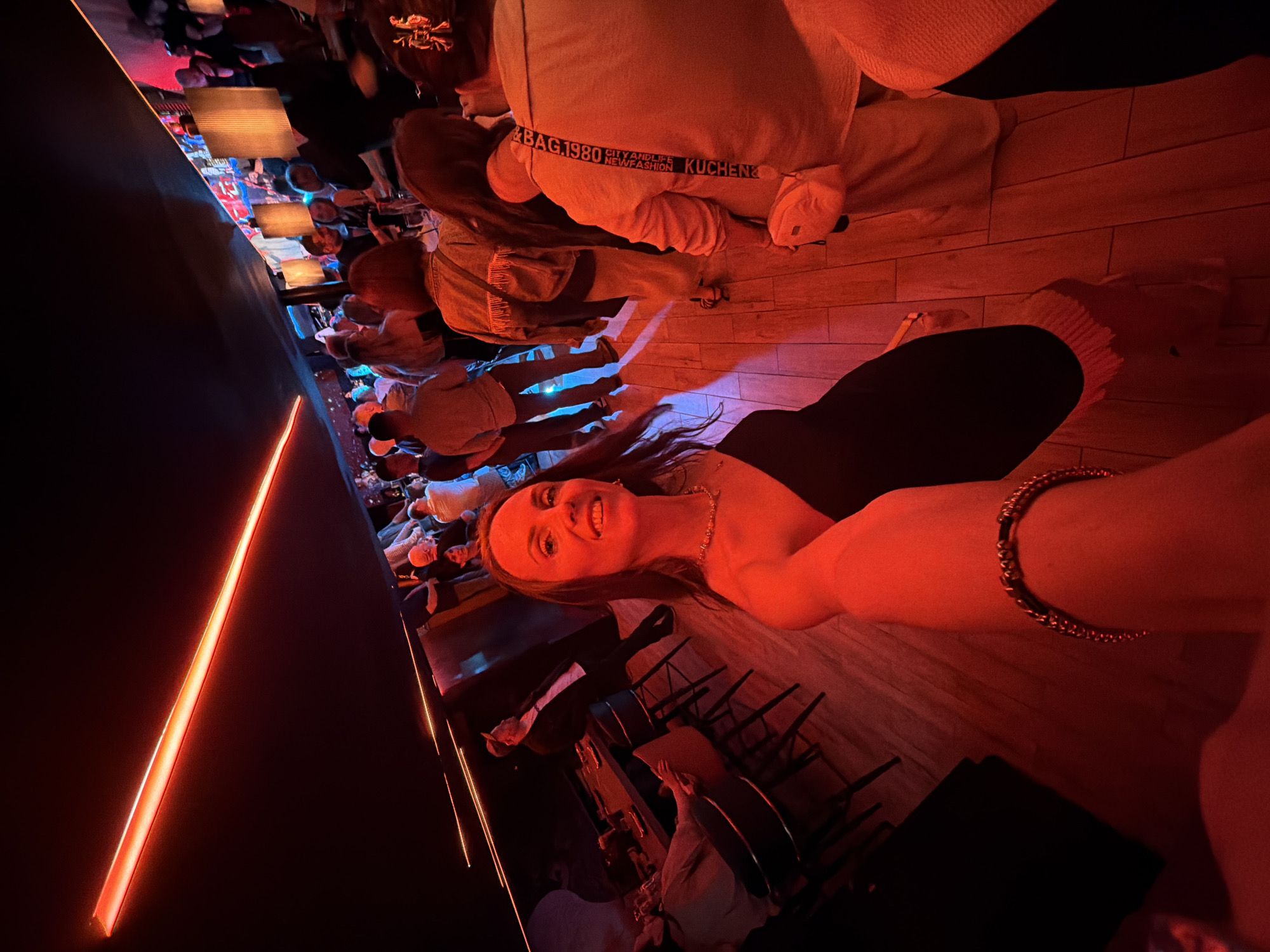 The image shows a group of people in what appears to be a dimly lit, modern establishment, possibly a bar or restaurant. The primary subject is a woman taking a selfie, smiling broadly, with a necklace and bracelet visible. Her hair is long and dark. She is wearing black pants and a top. The setting is characterized by warm, red-toned lighting, highlighting wooden floors and what seem to be tables and chairs in the background. Several other individuals are present, some seated and some standing, their faces mostly obscured by the lighting and angle.
A prominent object in the background is a light-colored garment, possibly a sweatshirt or blanket, with visible text that reads "BAG 1980 CITYANDLIFE NEWFASHION KUCHEN".
The atmosphere suggests a social gathering or outing, likely in the evening due to the low light conditions and artificial lighting. The woman's expression conveys happiness and enjoyment. The location context of Khabarovsk, Russia, is provided, but there are no specific visual cues within the image to confirm this location.