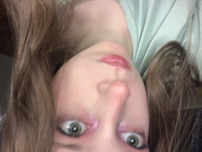A selfie-style image shows a person with pale skin, green eyes, and light brown hair. The image is rotated, so the face appears upside down. Pink eyeshadow is applied around the eyes. Lips are glossy and pink. A light green fabric is visible in the background. The subject's face is the primary focus.