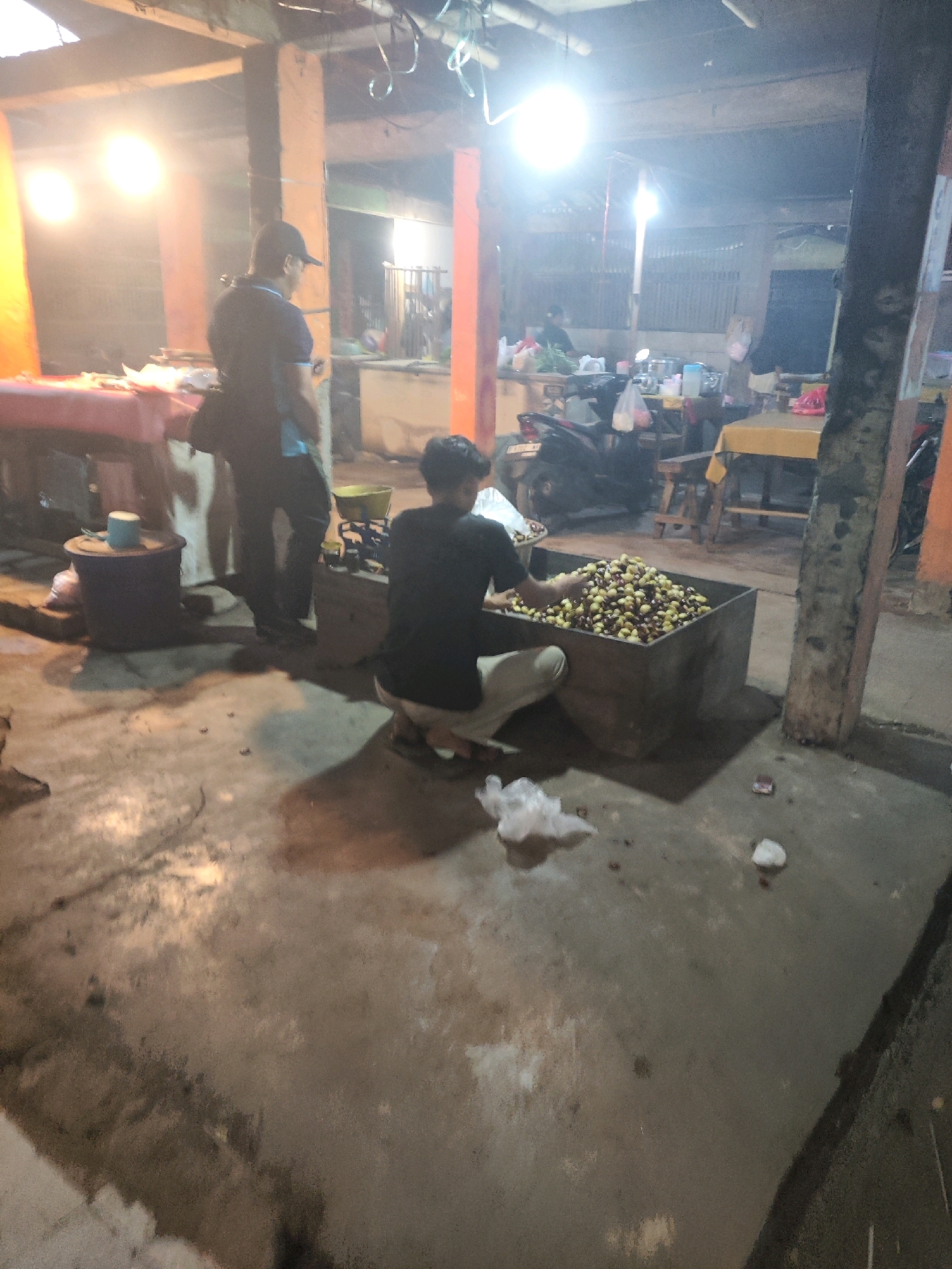 The image depicts an outdoor scene at night in a market area, possibly in Karawang, Indonesia, with bright overhead lighting. In the foreground, a young person wearing a black t-shirt and light-colored pants is crouched down, interacting with a large, rectangular metal container filled with a large quantity of small, round, brown and yellowish objects, likely produce. The person is reaching into the container with their right hand, appearing to sort or select the items. To the left, a person wearing a dark t-shirt and a baseball cap stands facing away from the camera, with a bag slung over their shoulder. Behind them, a red table is visible with some items on it. Further back, there are stalls with various goods, some tables, chairs, and a parked motorcycle. The ground is concrete, with some scattered debris and reflections from the lighting. The overall environment suggests a busy marketplace at the end of the day or during evening hours.