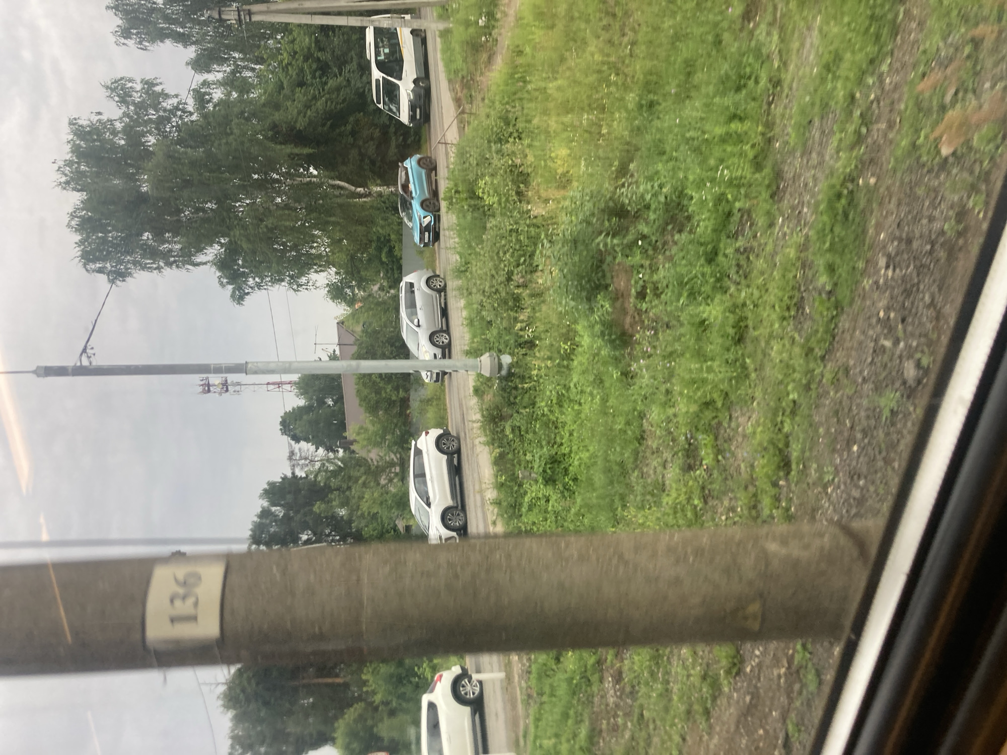 A street scene is captured from a window, showing a number of cars parked and moving along a road. In the foreground, a thick, textured concrete pole dominates the left side of the frame, with a small, light-colored plaque bearing the number "136" attached to it. The pole is out of focus, suggesting the photo was taken from a moving vehicle.
Beyond the pole, a paved road runs horizontally across the middle ground. Several cars are visible on the road. On the left, a white SUV is parked facing right. Further down the road, a white crossover SUV is in motion, also facing right. Next to it, a bright blue, sporty-looking car is driving. On the far right, a white van with yellow trim is driving away from the camera.
The environment is lush and green, with abundant vegetation lining the road. Tall trees with full foliage form a backdrop on either side of the road. In the distance, a cell tower with red and white sections can be seen rising above the trees, and a few residential buildings with peaked roofs are scattered among the greenery. The sky is overcast and appears cloudy, suggesting a temperate climate. The presence of the number "136" on the pole and the context of rural Russia suggest a location in the village of Sokolovo, Russia, possibly a section or route number.