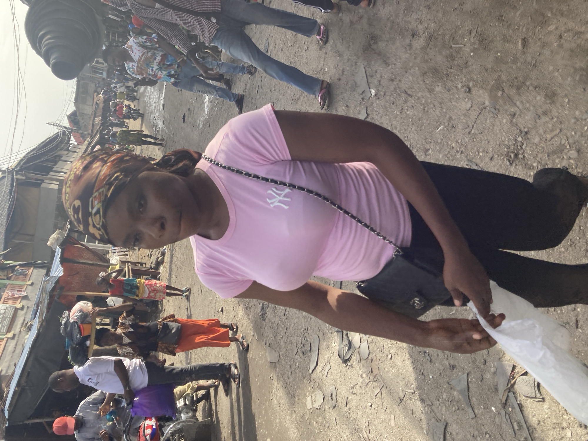 The image shows a woman in a pink t-shirt and black pants holding a white plastic bag in a street in Ura Nta Umuarandu, Nigeria. She is wearing a patterned headscarf and a crossbody bag. The street appears to be a bustling market area, with other people, motorcycles, and structures visible in the background. The ground is uneven, covered with debris.