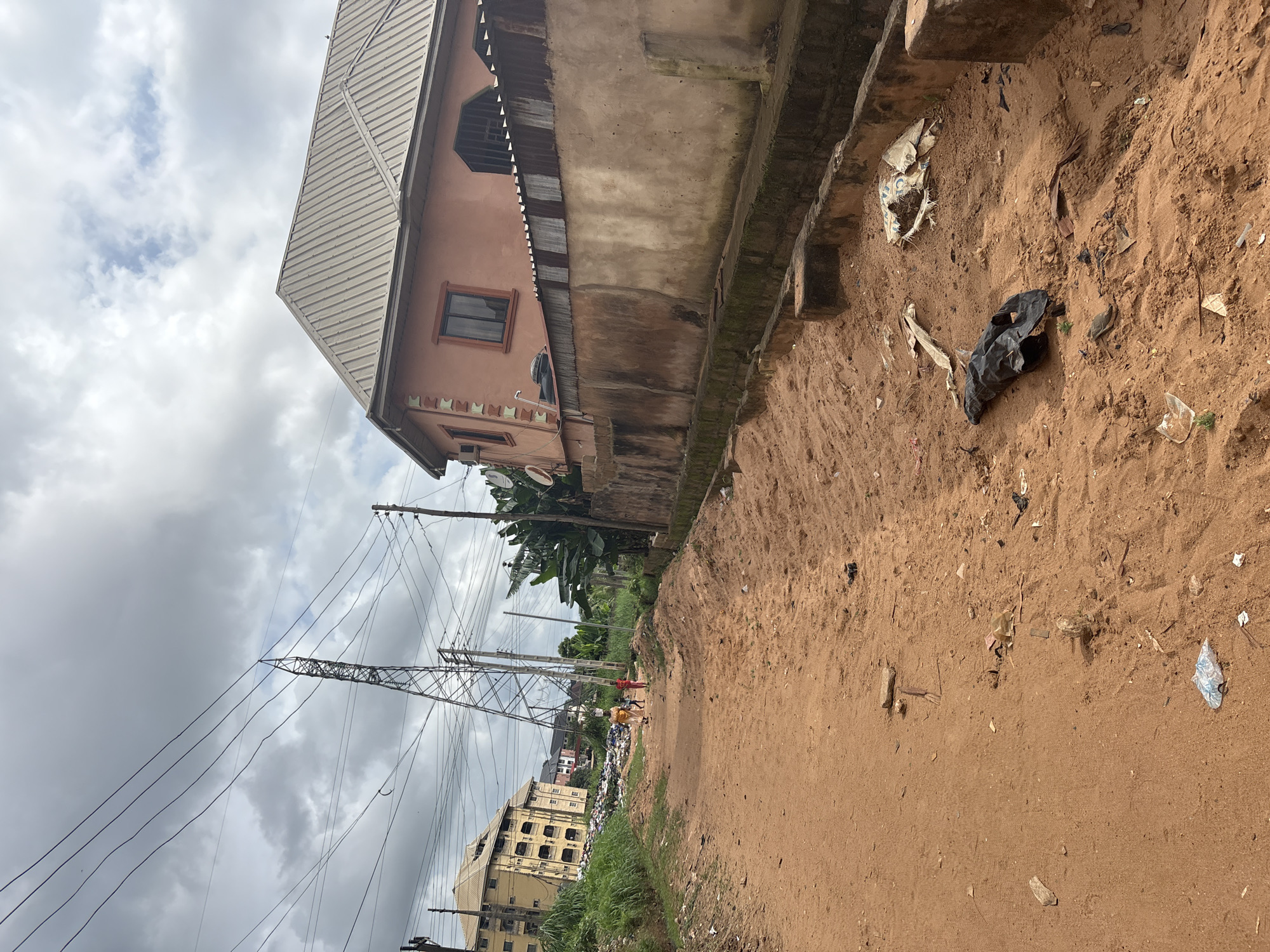 A dirt road in Nkpor, Nigeria, is lined with a drainage ditch on the right and a pile of refuse on the left. A two-story building with an orange facade and a metal roof is visible on the right side of the road. Electrical poles and wires stretch across the sky. Three people are walking down the dirt road in the distance, and a large metal tower is visible behind them. The ground is covered in sand and scattered with litter, including plastic bags and debris. The sky is overcast with grey clouds.