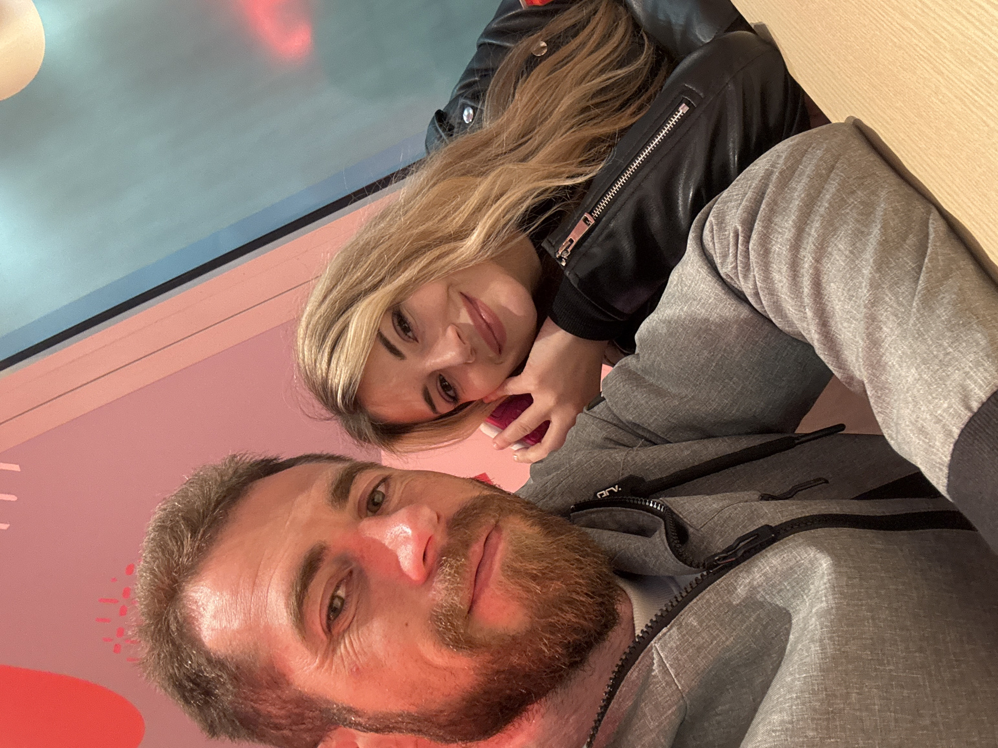 A selfie, slightly rotated counter-clockwise, shows two individuals, a man and a woman, indoors. The man, in the foreground, has short hair, a beard, and is wearing a grey zip-up jacket. The woman, positioned behind and to the side of the man, has long, blonde hair and wears a black leather jacket. She holds a pink mobile phone. The background consists of a pink wall with red and white abstract designs and a light blue ceiling. The photograph was taken in Budva, Montenegro.