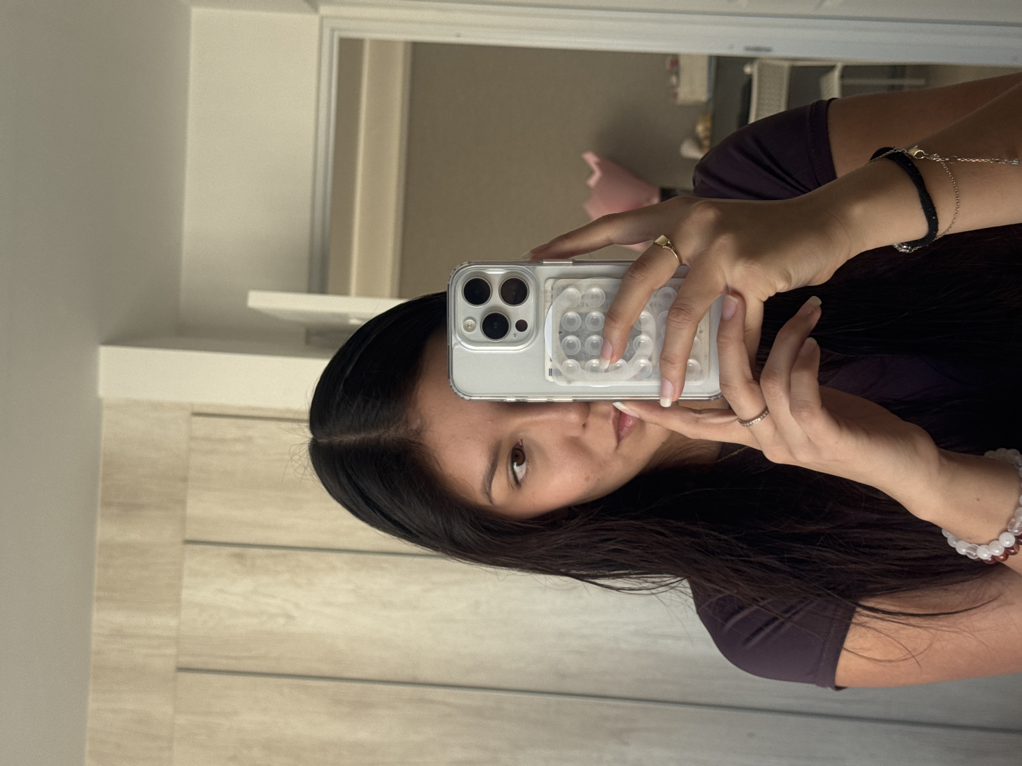 A woman is taking a selfie in front of a mirror. She is holding a phone with a clear case that has a white, circular pattern on the back. She has long, dark hair. Her face is partially obscured by the phone. She is wearing a purple shirt. There are bracelets on her right wrist and rings on her fingers. The background includes a white door frame and a glimpse of another room in the mirror. The location is not San Francisco Ocotlán (Ocotlán), Mexico.