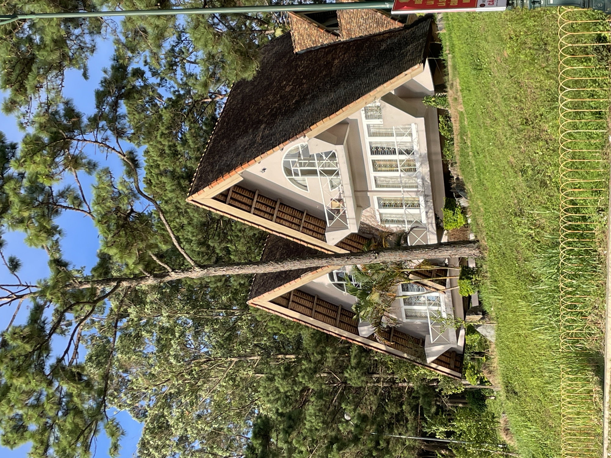 Two A-frame houses with brown tile roofs are situated on a grassy hillside. The houses are light-colored with multiple windows and balconies. Tall evergreen trees surround the houses, and a yellow metal fence is visible in the foreground. A red and white sign is partially visible on the right side of the image. The setting appears to be a natural, outdoor environment, likely in a mountainous or hilly area within or near Da Lat, Vietnam, a city known for its pine forests and cooler climate.