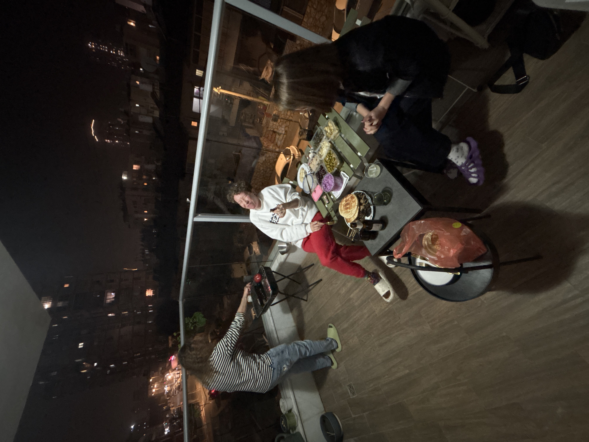 The image shows a group of three people on a balcony in Bat Yam, Israel, at night. The cityscape is visible in the background with numerous buildings. One person wearing a striped shirt and blue jeans is tending to a small barbecue grill. A second person, wearing a white sweatshirt and red pants, is sitting at a table. A third person wearing dark clothing is seated at the same table. The table has a spread of food and drinks. Other objects present include a chair, a grill, plates, and cutlery.