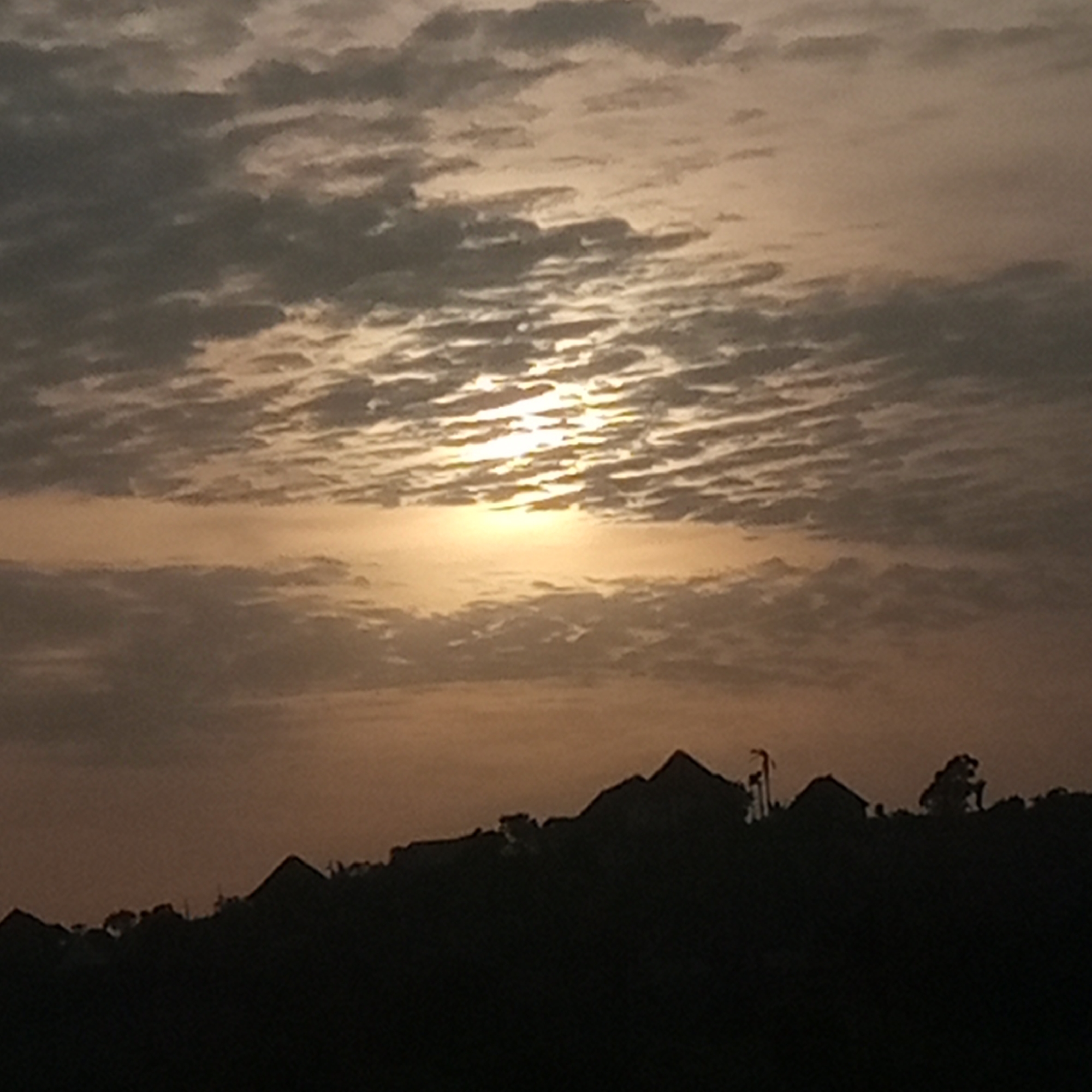 The image depicts a sunset or sunrise over a silhouette of buildings and trees. The sky is filled with textured clouds, partially obscuring a bright, golden sun. The foreground shows dark shapes of what appears to be residential or rural structures interspersed with trees. The location and city are indiscernible. There are no observable people, actions, or interactions.