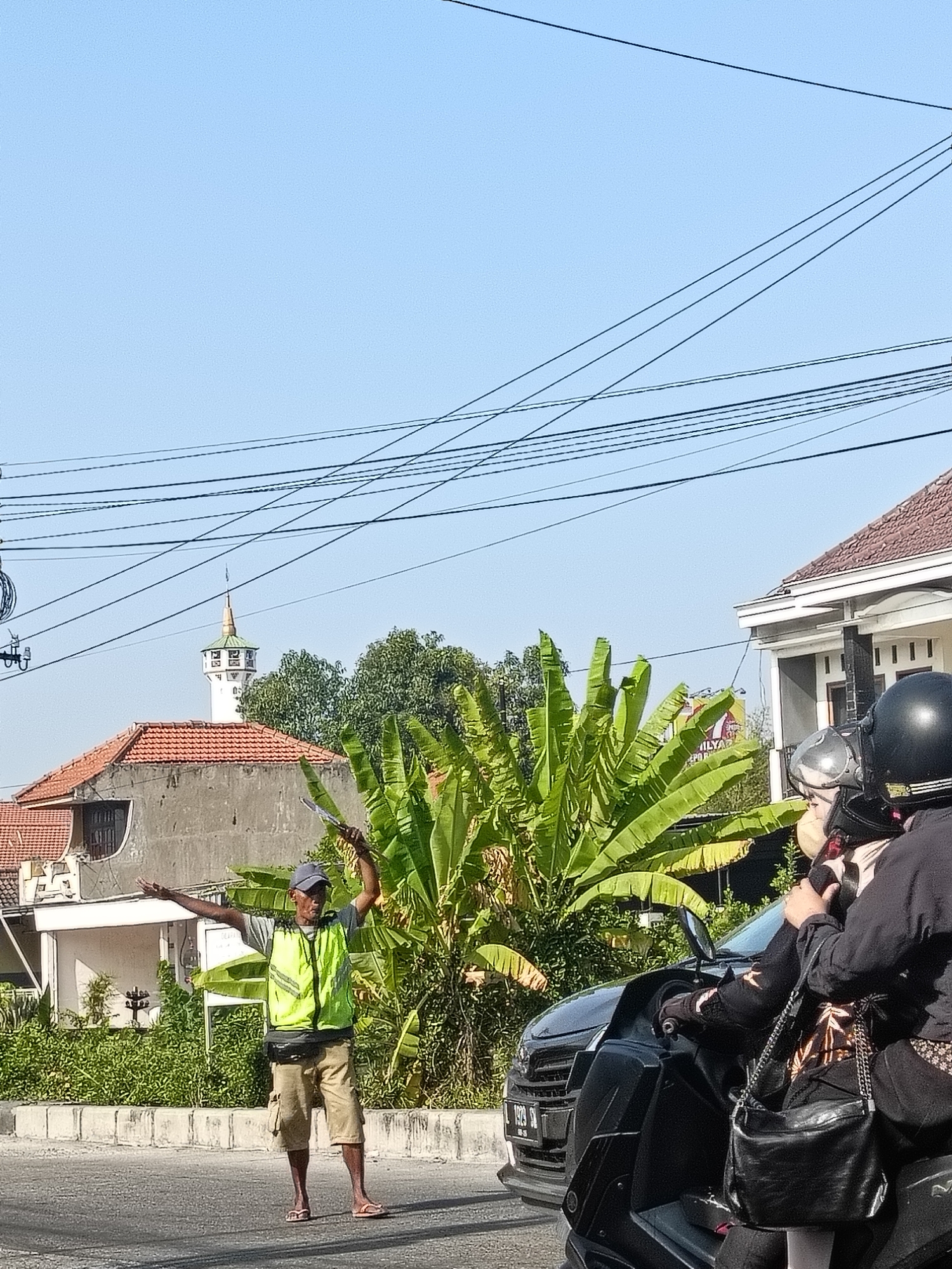 A traffic director wearing a yellow-green reflective vest and a grey baseball cap stands in a street in Gedangan, Indonesia, gesturing with his arms. He is holding a piece of paper in his raised left hand. He wears tan shorts, a grey t-shirt, and flip-flops. A black fanny pack is around his waist. A motor scooter carrying two individuals is stopped next to him in the foreground. The person in the back is wearing a black hijab and a patterned headscarf, and the person in front is wearing a helmet and a black jacket. A black purse is hanging from the shoulder of the person in the back. In the background, a mosque with a tall white minaret and a gold-tipped roof is visible behind a cluster of green trees. There are power lines overhead, and houses with terracotta roofs are also present. The scene is illuminated by sunlight under a clear blue sky.