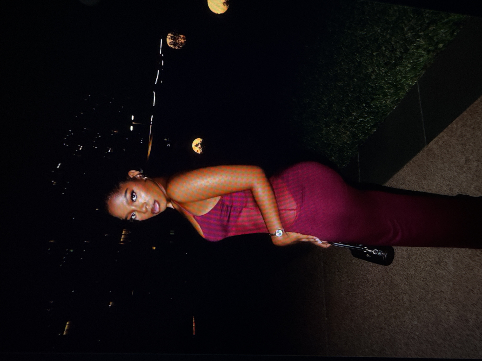 A woman is posing in a tight, sleeveless, deep red dress with sheer panels on the sides. She is wearing a silver wristwatch on her left wrist and is holding a black handbag with a silver clasp in her left hand. Her hair is styled in an updo, and she is wearing makeup. The background is dark, with out-of-focus lights from buildings and several round, glowing lights, possibly decorative lamps, scattered in the distance. A patch of green grass is visible to the right, bordered by dark pavement. The scene appears to be outdoors in an urban setting at night, potentially in Abuja, Nigeria, given the context.