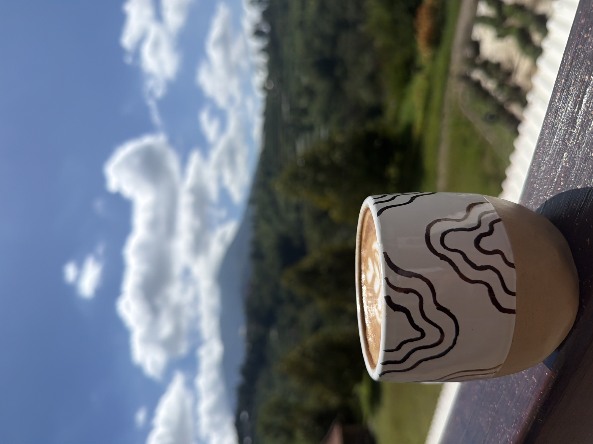 A close-up shot focuses on a latte in a decorative ceramic mug, placed on a wooden railing. The mug has a white base adorned with abstract brown wavy lines and a beige bottom. The latte art on the coffee surface features a delicate pattern. In the background, a blurred landscape unfolds with rolling green hills, lush trees, and a majestic mountain under a bright blue sky dotted with fluffy white clouds. The scene is outdoors, suggesting a tranquil setting with a scenic view. The location is identified as Baturiti, Indonesia.
