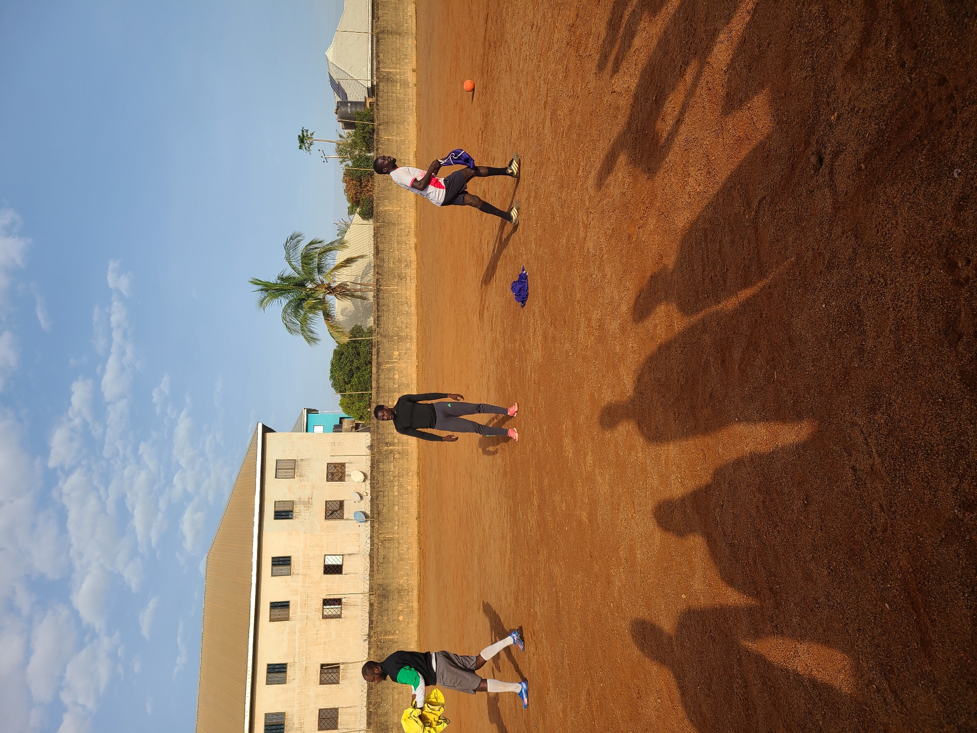 In Barakin Lerawi, Nigeria, three men are present on a dirt field under a clear blue sky with sparse clouds. A light-colored building with numerous windows is visible in the background, behind a low, rough-textured wall. A palm tree stands to the right of the building, and other trees and structures are further in the distance. One man, on the left, wears a green and black long-sleeved shirt, gray shorts, white socks, and blue athletic shoes. He is carrying a bundle of yellow fabric. In the center, a man in a black long-sleeved shirt and dark pants with pink athletic shoes stands facing right. On the right, a man in a white and red shirt, black shorts, and black and white athletic shoes is looking to his left, holding blue fabric. An orange ball is on the ground to his right, and a blue cloth is on the ground to his left. Long shadows are cast across the field from the men and likely other unseen individuals.