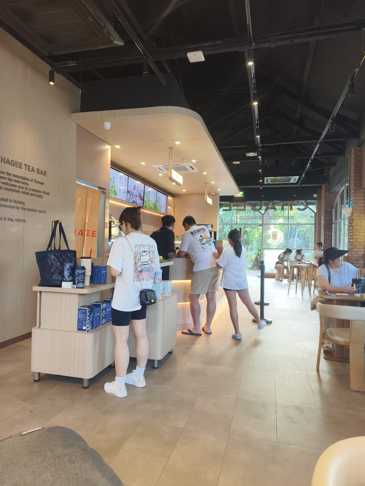 The image depicts the interior of a modern tea bar, identified as CHAGEE, in Kuta Selatan, Indonesia. Several individuals are present, with some ordering or waiting at the counter, while others are seated at tables. The decor features a light wood finish on the counter and walls, complemented by dark ceiling beams and industrial-style lighting. Merchandise, such as tote bags and packaged tea, is displayed on shelves near the entrance. Large windows overlook a lush green outdoor area, suggesting a tropical setting. A digital menu board displays various drinks and promotions. The overall atmosphere is relaxed and contemporary, typical of a popular cafe or beverage establishment.