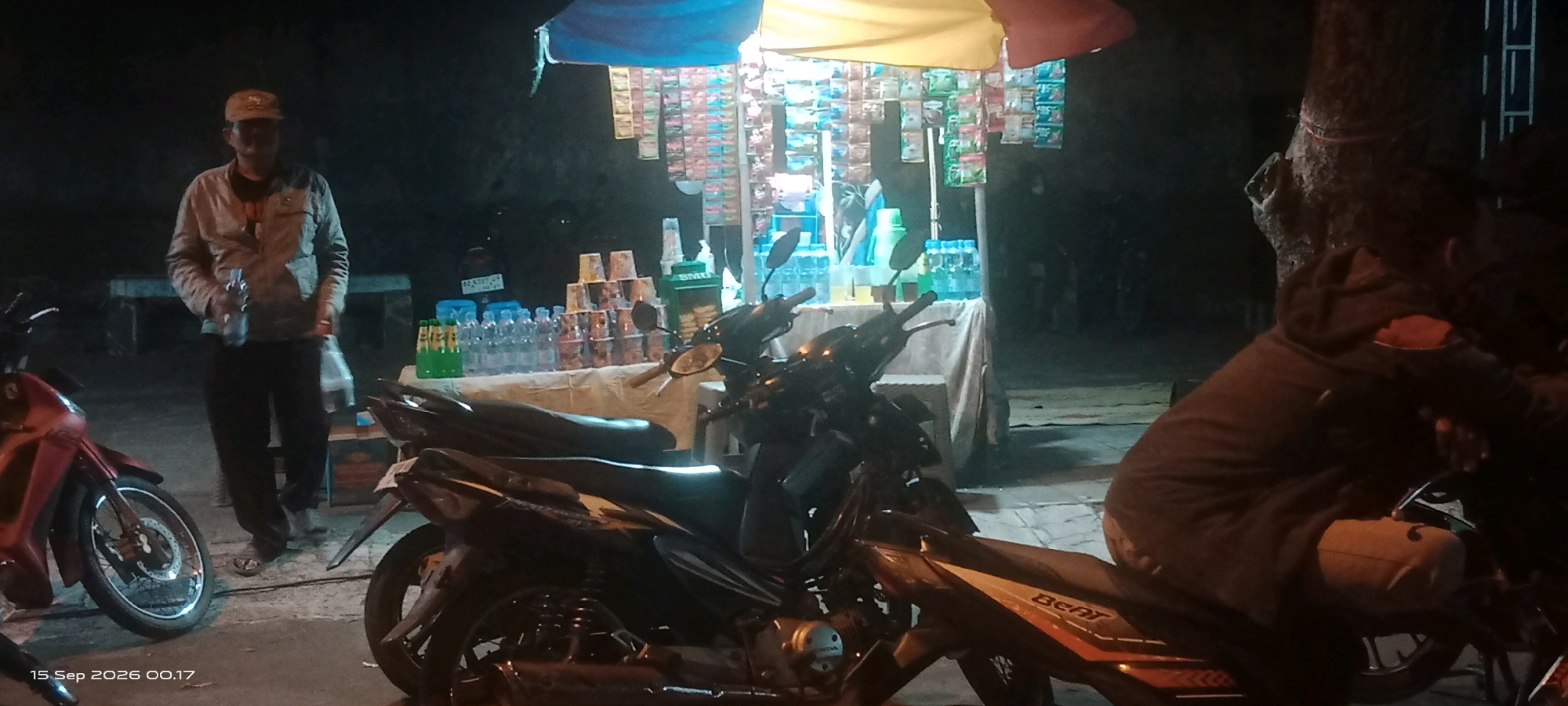 A man wearing a light-colored jacket and a cap stands near a stall selling drinks and snacks. The stall is decorated with colorful flags and displays various bottles of beverages and stacked cups. Several motorcycles are parked in front of the stall, suggesting a roadside or parking area. The scene appears to be at night, as indicated by the artificial lighting and the dark surroundings. The timestamp "15 Sep 2026 00.17" is visible in the lower left corner, indicating the date and time the image was captured.