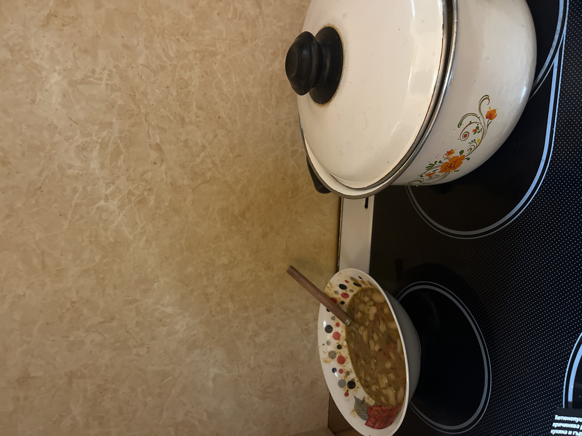 The image shows a kitchen setting, likely indoors. On the right side of the frame, there is a black stovetop with two circular heating elements, one of which has a white pot placed on it, covered with a lid that has a black knob. To the left and slightly below the pot, there is a white plate with a patterned rim containing what appears to be a stew or bean-based dish with a wooden spoon resting in it. The background on the left side of the image is a light tan wall, possibly with a marble or textured finish. Based on the text at the bottom right of the hob, it can be inferred that the location is likely in Burgas, Bulgaria. There are no people visible in the image.