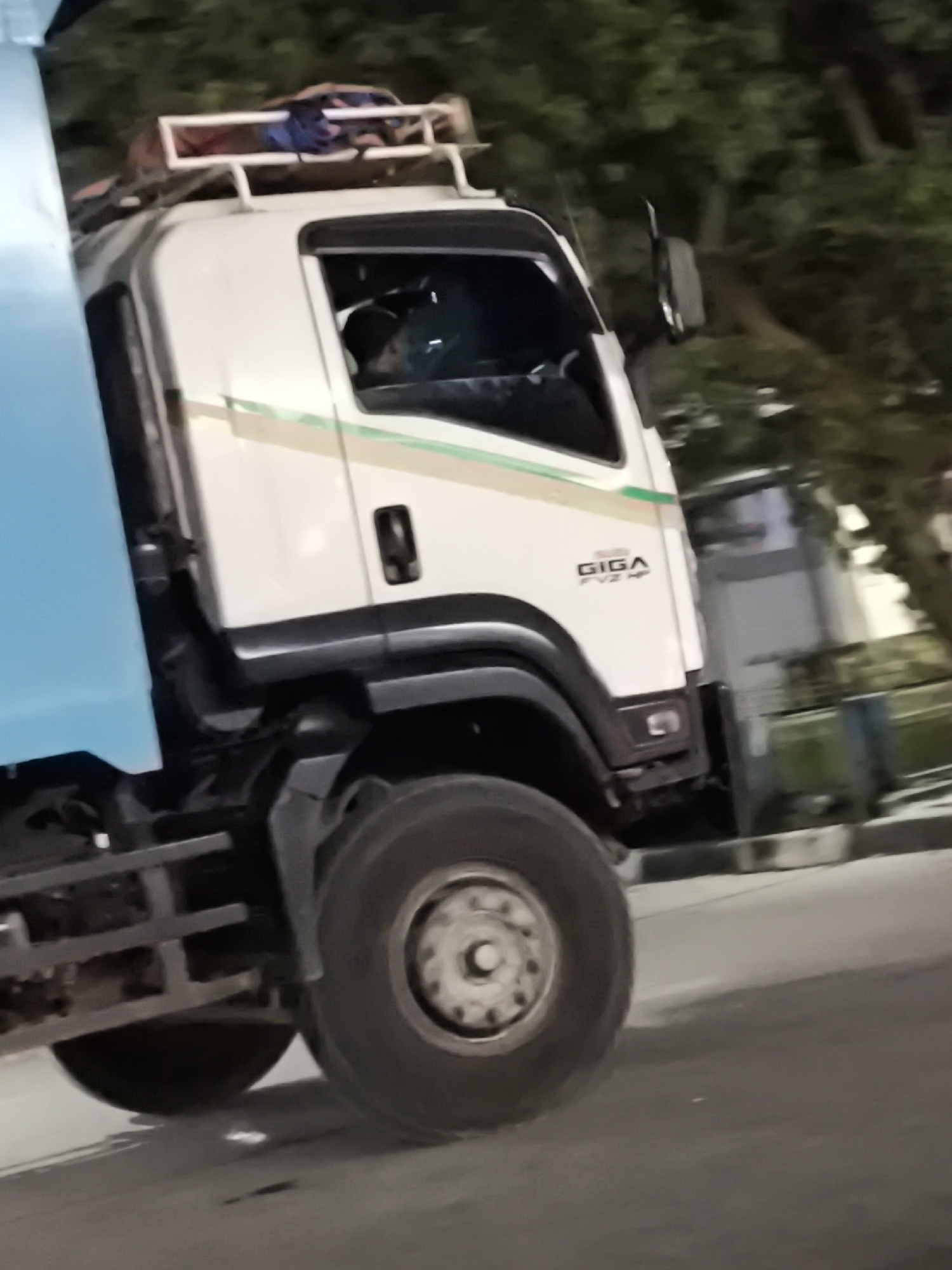 The image shows the side view of a white Isuzu GIGA FVZ HP truck, viewed from the front right, at night in Waringinkurung, Indonesia. The truck has a blue cargo bed on the left side and a roof rack carrying bags on top of the cab. The driver, a person with dark hair, is visible inside the cab. The truck's large front tire is prominent in the foreground. The background consists of out-of-focus trees and a building. The scene appears to be on a street or road.