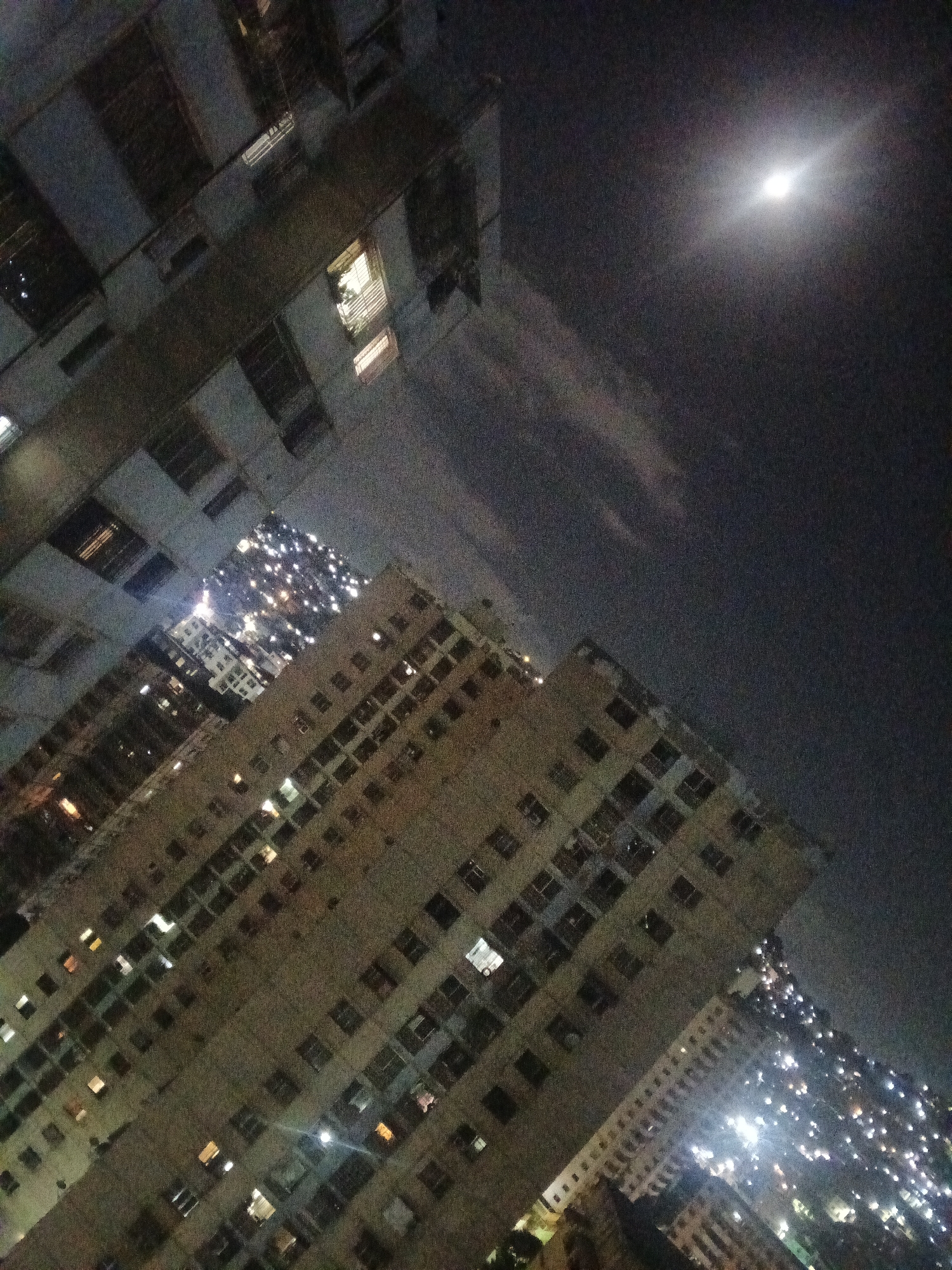 The image shows a nighttime view of buildings in Caracas, Venezuela. The buildings are tall, multi-story apartment complexes. Many windows are illuminated, suggesting the presence of people inside. In the upper right corner, a bright moon shines through the night sky. The scene evokes a sense of urban density and night life in the city.