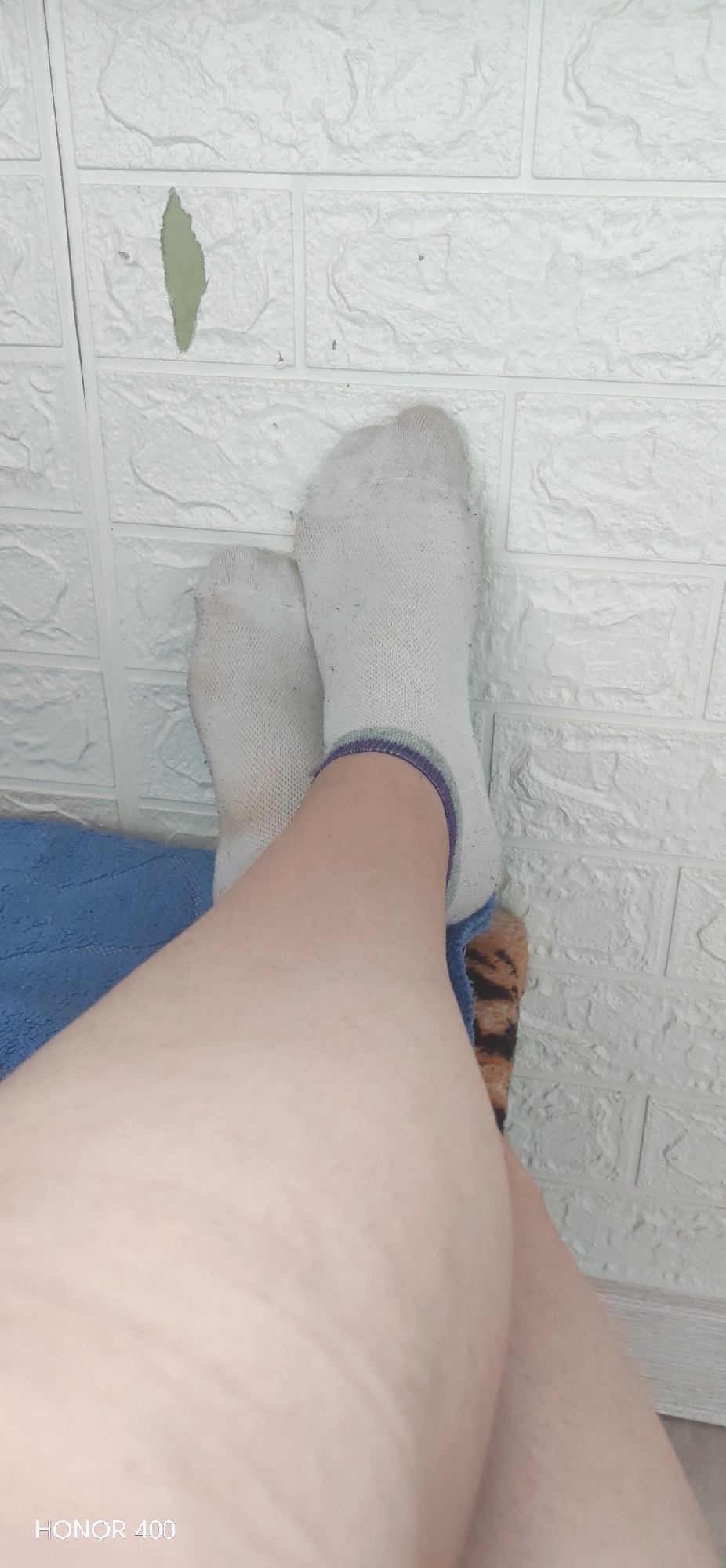 The image shows a pair of legs with white socks against a textured white brick wall. The socks have striped trim at the ankles. The legs are positioned on what appears to be a blue cloth and a material with a tiger stripe pattern. A partially peeled section is visible on the brick wall. The bottom left corner of the image contains the text "HONOR 400." There is no evidence to suggest the location is in the city of Noviy, Russia.
