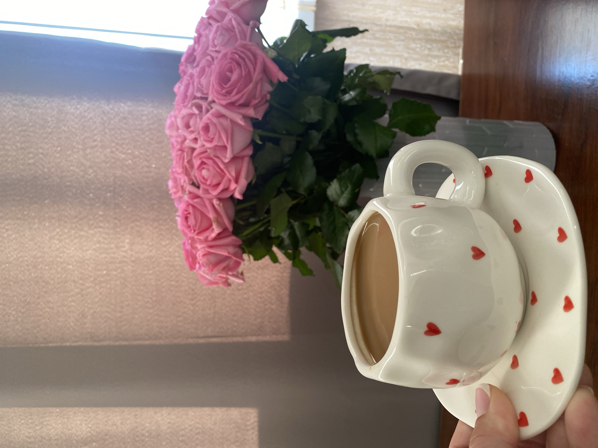 A vase filled with pink roses is positioned on a dark brown table, next to a white teacup and saucer adorned with red hearts. The teacup is partially filled with a beige-colored liquid, suggesting tea or coffee. The scene is illuminated by soft, diffused light filtering through a window, as seen from the shadow on the left of the image, casting a gentle glow across the textured wall behind the roses. A hand holding the saucer is on the right edge of the image. The setting suggests an indoor location, possibly a home or café, in Sochi, Russia.
