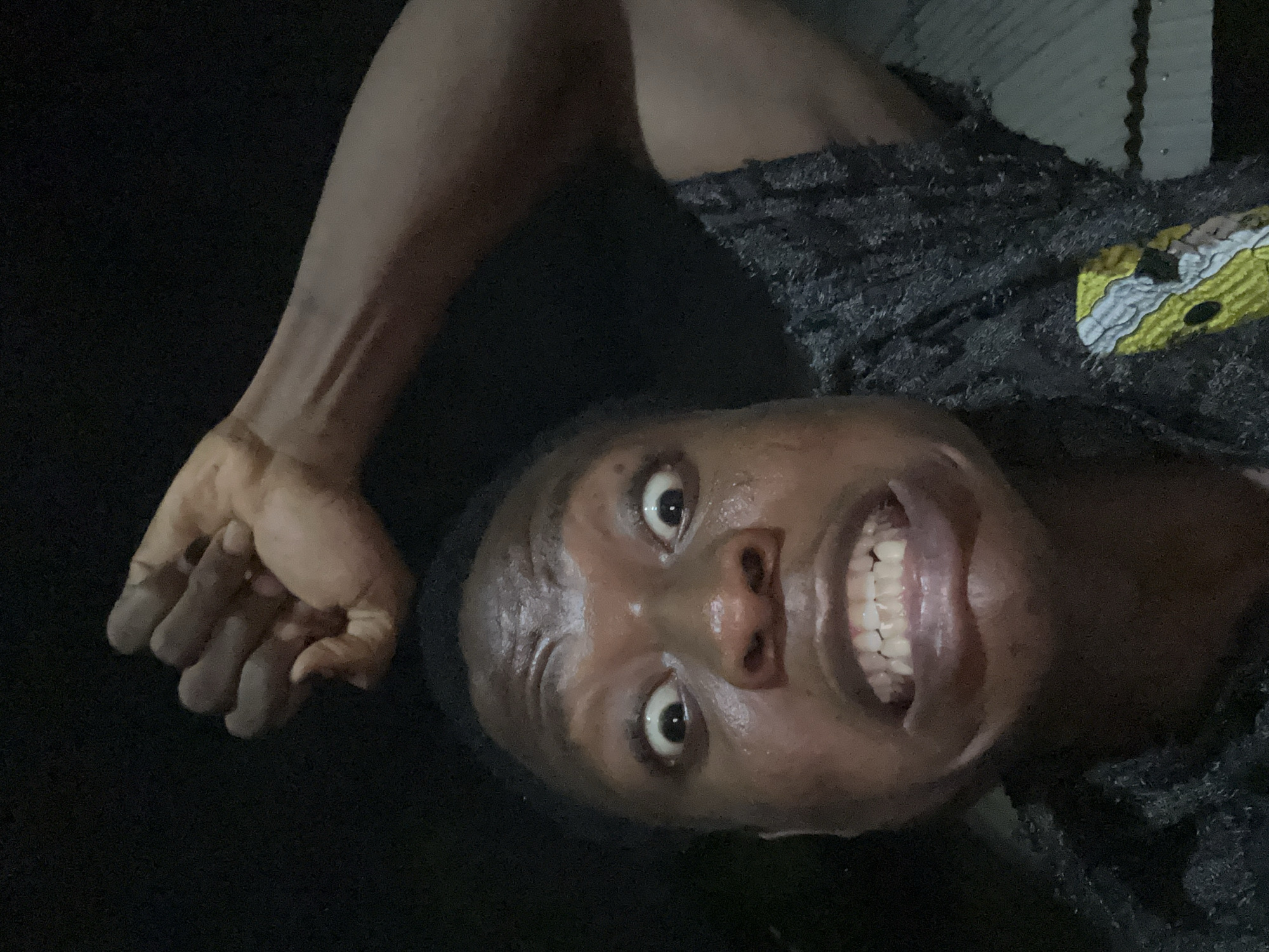 A person is positioned at an angle in a dark environment. The individual is smiling widely, exposing their teeth. Their face appears slightly sweaty. The person's left arm is raised. The location is unidentified.