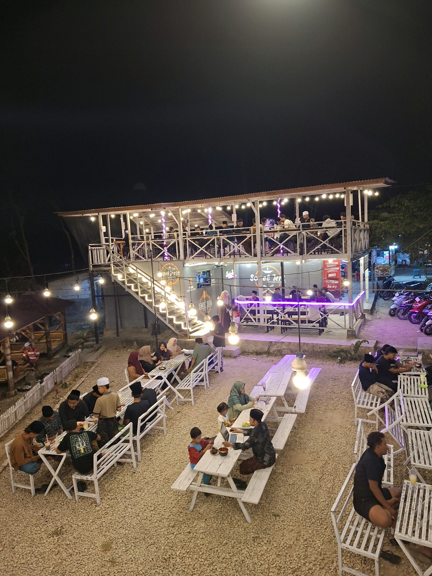 This is an elevated, nighttime, wide-angle shot of an outdoor dining area in Lantek Barat, Indonesia, illuminated by string lights and a few larger bulbs. The establishment appears to be a multi-level building with a corrugated metal roof, featuring an outdoor staircase leading to an upper level. Several tables and benches are arranged on a gravel ground. People are seated at various tables, some appearing to be dining, while others are socializing. The scene is populated by individuals, predominantly males, some wearing traditional Islamic attire like peci (caps). The environment suggests a casual, communal dining experience, possibly a cafe or restaurant. Motorcycles are parked in the background, indicating a street-side or accessible location. The overall atmosphere is lively and social, with the artificial lighting creating a warm glow against the dark sky.