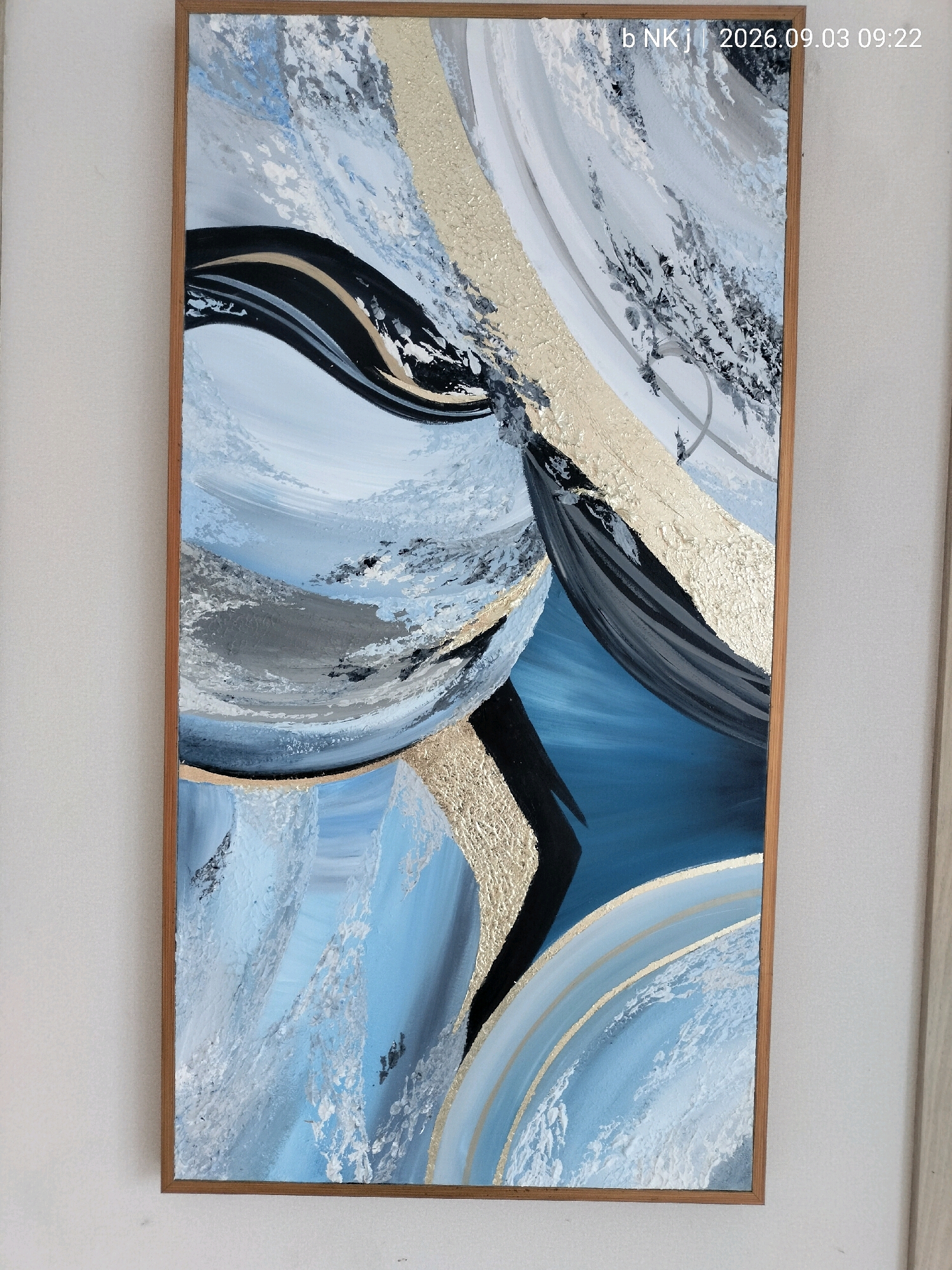 A vertical, abstract painting with a wooden frame hangs on a white wall. The painting features a dynamic composition of swirling shapes and textures in shades of blue, white, black, grey, and gold. The colors are applied with varying techniques, including thick impasto strokes and smooth washes. The gold elements appear to be textured, possibly using gold leaf or textured paint. The overall impression is one of movement and depth, with a sense of flowing forms. The top right corner of the painting displays a watermark with the text "b NKJ | 2026.09.03 09:22", indicating the date and time the image was captured. The background wall is textured and shows faint vertical lines, suggesting it might be a textured wallpaper or painted surface. The lighting in the scene appears to be soft and diffused, casting minimal shadows. The image is a close-up shot, focusing on the painting and its immediate surroundings. There are no people or identifiable landmarks visible in the image.