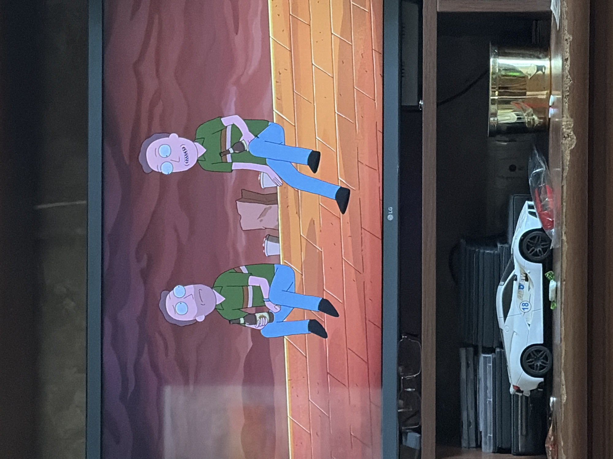 Two male cartoon characters are seated on a brick wall. Both characters wear green shirts with horizontal stripes and blue pants. The character on the left holds a bottle in his right hand. The character on the right also holds a bottle in his left hand. Behind them, a sandy environment is depicted with some bags and containers. The scene is displayed on a television screen. Below the television, a shelf holds various items including a white toy car, DVDs, glasses, and a gold cylindrical object.