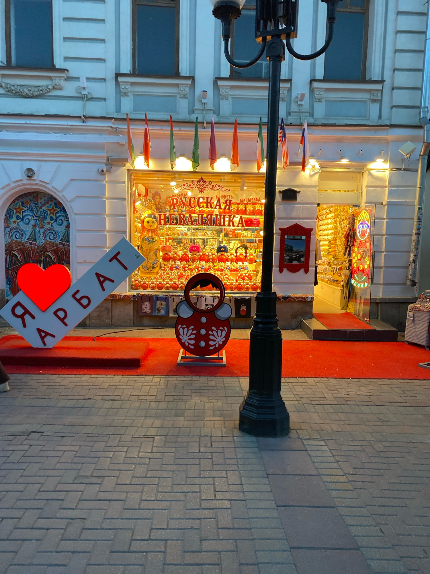 The image shows a street scene in Moscow, Russia, specifically on Arbat Street. In the foreground, a red carpet leads up to a storefront displaying Russian nesting dolls and souvenirs. A large, illuminated red heart is attached to a tilted sign that reads "Я АРБАТ" (I Arbat) in red letters. A cutout of a matryoshka doll, painted red and white, stands on the carpet. In the background, the facade of the building features arched windows and architectural details, with several flags displayed above the storefront. A lamppost stands in the middle of the street, partially obscuring the view of the building. The street is paved with cobblestones. The lighting suggests it is either dusk or early evening.