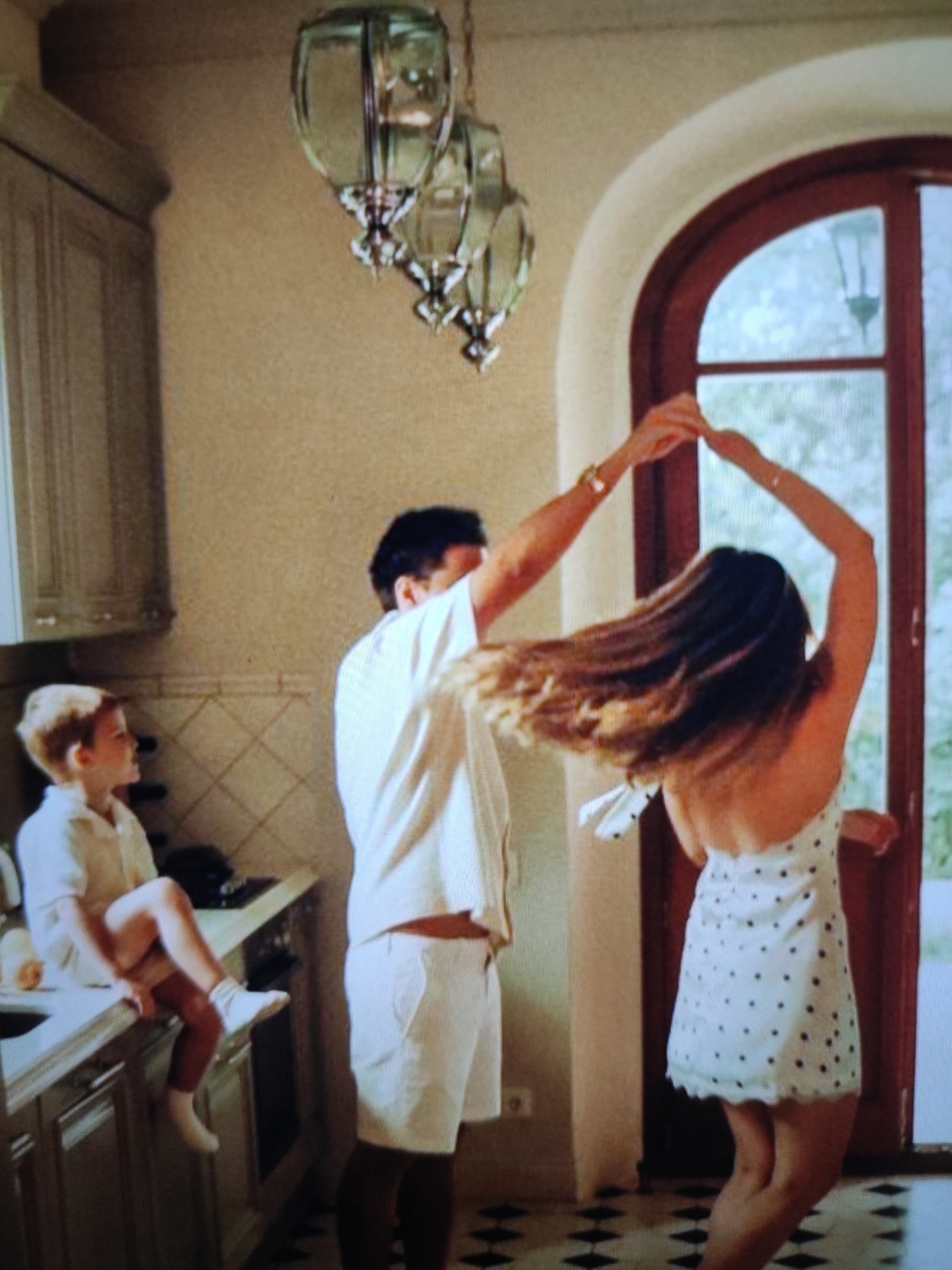 The image captures an indoor scene, possibly a kitchen or dining area, featuring three people. In the foreground, a woman with long, dark hair is dancing, her back to the viewer, wearing a white polka-dot dress. She is holding hands with a man who is facing away from the camera, dressed in a white shirt and shorts. A young boy with blond hair sits on a countertop to the left, wearing a light-colored shirt and shorts, and white socks. The background shows a large arched doorway, a chandelier with three globes hanging from the ceiling, and kitchen cabinetry. The floor has a black and white diamond pattern. The lighting is warm and ambient.