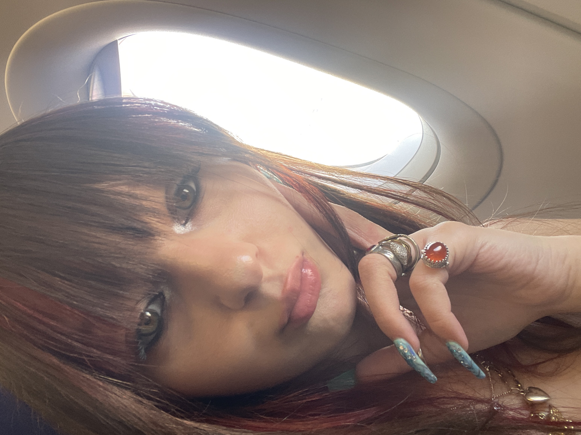 This is a close-up, slightly blurred, selfie-style photograph of a young woman with long, dark brown hair with reddish highlights, posing next to an airplane window. The woman is looking towards the camera with her head tilted slightly. She is wearing makeup, including eyeliner and mascara, and has glossy pink lipstick on her lips. She has long, pointed fingernails painted a light blue with glitter. Her right hand is raised, with several rings on her fingers, including one with a large orange stone and another with multiple bands. She is wearing a necklace with a heart-shaped pendant. The background shows the bright interior of an airplane cabin, with the curved edge of the window frame visible on the right side of the image. The lighting suggests it is daytime, with bright light coming from outside the window. The image does not contain any visual elements that would indicate a specific location in the city of Yagel, Israel.