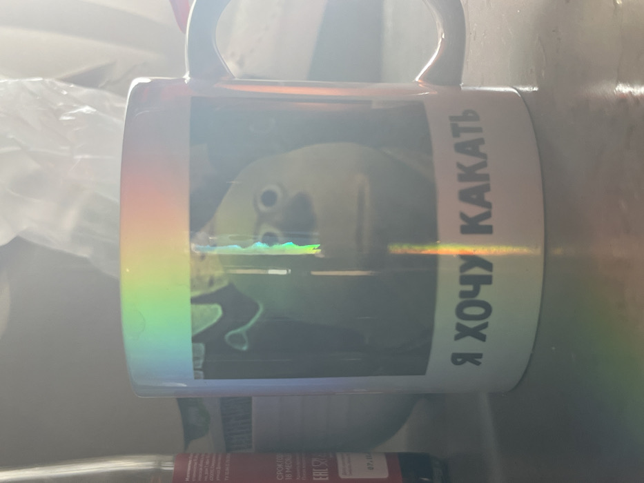 A cylindrical mug features a gradient from green to red on its left side. A mirrored image of a cartoon face is visible, partially obscured by reflections. The right side of the mug is white, printed with Cyrillic text "Я ХОЧУ КАКАТЬ" in black. The mug has a handle and is positioned on a surface amidst miscellaneous items, including what appears to be bottled sauce. Bright sunlight casts a strong glare across the surface of the mug.