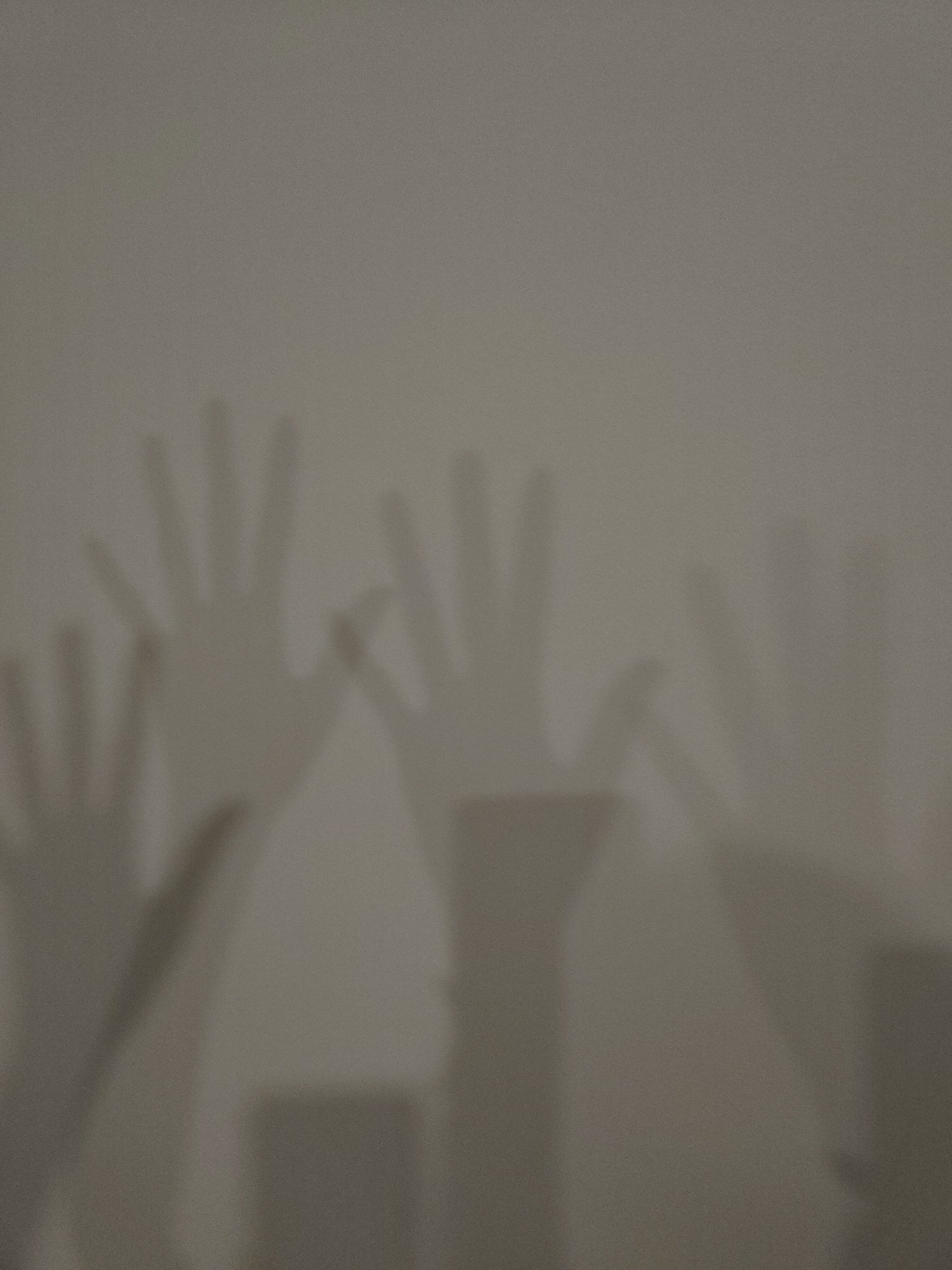 The image displays a series of hand shadows projected onto a plain, light-colored wall. Several human hands are visible as dark silhouettes against the lighter background, appearing to reach upwards or wave. At least four distinct hand shapes can be identified, with some fingers clearly splayed and others partially overlapping or less defined. The shadows are soft, indicating a diffused light source, and the wall itself has a slightly grainy texture. The scene depicts an activity of creating hand shadows, likely playful or artistic in nature, suggesting human interaction with light and surface. No specific objects other than the wall are visible. The setting is indoors, with no discernible architectural features or decorative elements providing further location cues. The time of day and weather cannot be determined. There is no visible text in the image.