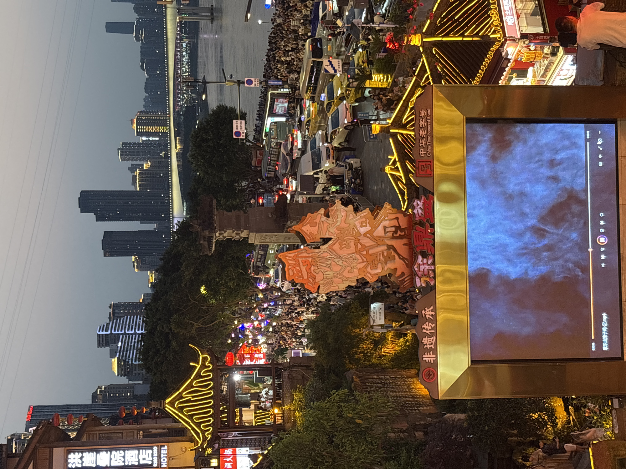 The photograph shows a street scene in Yuzhong District, China at dusk, featuring a large crowd gathered along a thoroughfare with visible traffic and buildings in the background. A waterway or river with a bridge is faintly visible in the upper portion of the image, juxtaposed with an urban skyline of high-rise buildings. Street lights and decorative lighting illuminate the area, highlighting traditional architectural elements and commercial establishments. A modern video display encased in an ornate frame is prominently featured, showing a video with playback controls visible on the screen. The camera angle and image orientation suggest the photograph was taken from an elevated perspective.