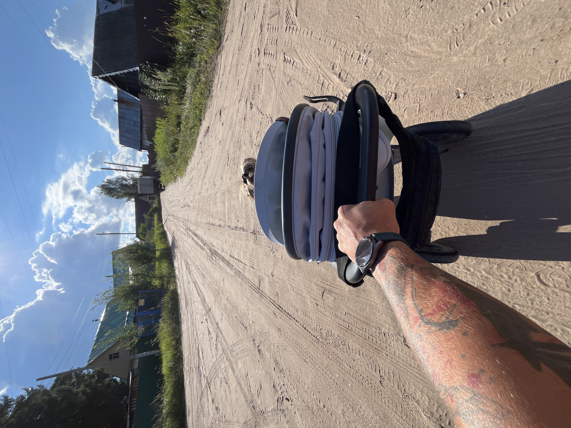 The image shows a first-person perspective of someone pushing a stroller on a dirt road in a rural area. The road is unpaved and has tire tracks visible. On either side of the road are houses with corrugated metal roofs and fences. Green grass and some trees are also present. The sky is blue with fluffy white clouds and the sun is shining brightly, casting shadows. A person's tattooed arm and hand are visible, holding the stroller handle. A watch is worn on the person's wrist. A child's hair is visible inside the stroller. The scene depicts a sunny day in what appears to be a village or small town, possibly in Russia.
