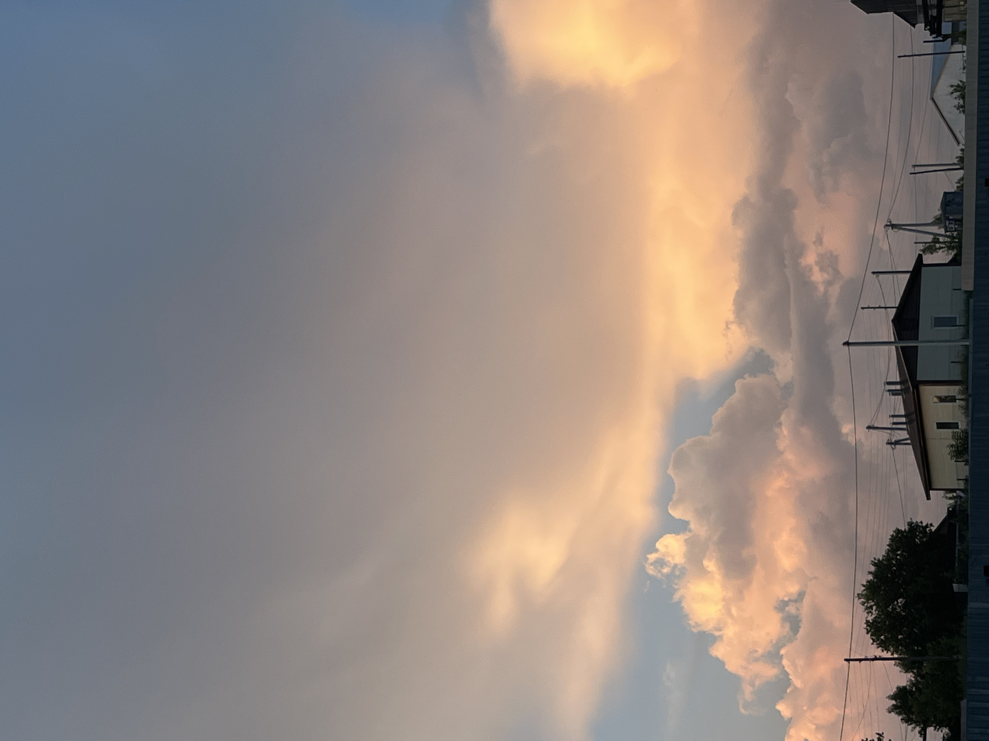 The image shows a twilight sky over the city of Ludorvay, Russia, with large, fluffy clouds illuminated by the setting sun, casting a warm, orange glow. The sky transitions from a soft blue at the top to a hazy, rosy hue towards the horizon. Silhouetted against the sky are a few simple, low-rise buildings with dark roofs, suggestive of a rural or suburban setting. Electrical wires stretch across the scene, supported by utility poles. To the left, a dense cluster of dark green trees stands out. A blue fence runs along the bottom of the frame, partially obscuring the base of the buildings and trees. The overall impression is one of a peaceful evening scene in a quiet locality.