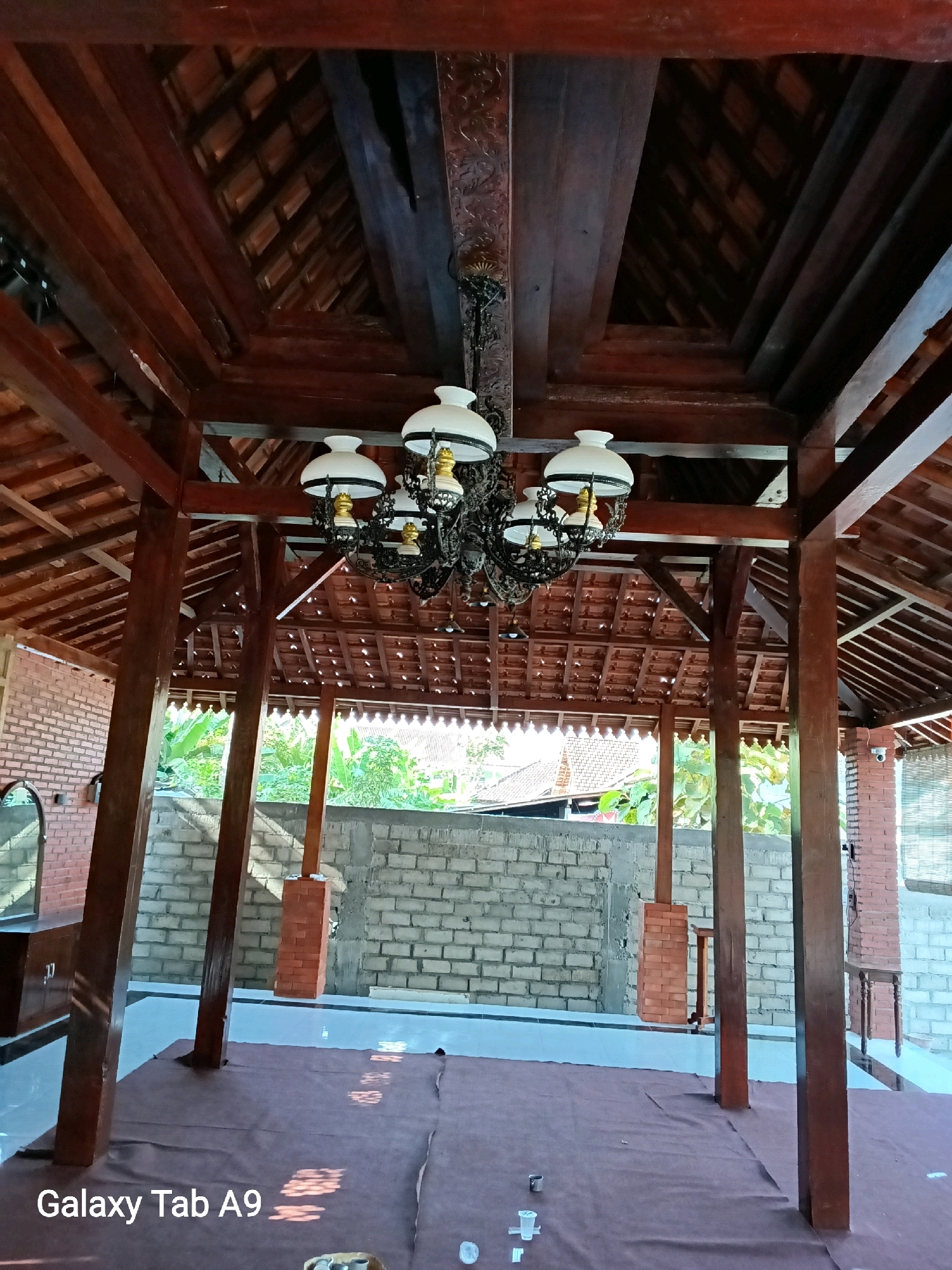 The image captures an interior view of a traditional Indonesian-style pavilion, likely located in Trenggalek, Indonesia, given the presence of the "Galaxy Tab A9" watermark, a common device used by local photographers. The pavilion features a prominent wooden roof structure with exposed beams and rafters, supporting terracotta roof tiles. A decorative, multi-bulb chandelier hangs from the center of the ceiling. The walls are constructed of light-colored bricks, with some sections of darker brickwork visible. In the foreground, a brown carpet covers the floor. Several wooden pillars support the roof. Through the open sides of the pavilion, lush green foliage and glimpses of buildings with red tile roofs can be seen, suggesting an outdoor or semi-outdoor setting. The lighting is natural and appears to be from daylight filtering into the space, creating dappled shadows on the floor. No people are visible in the scene.