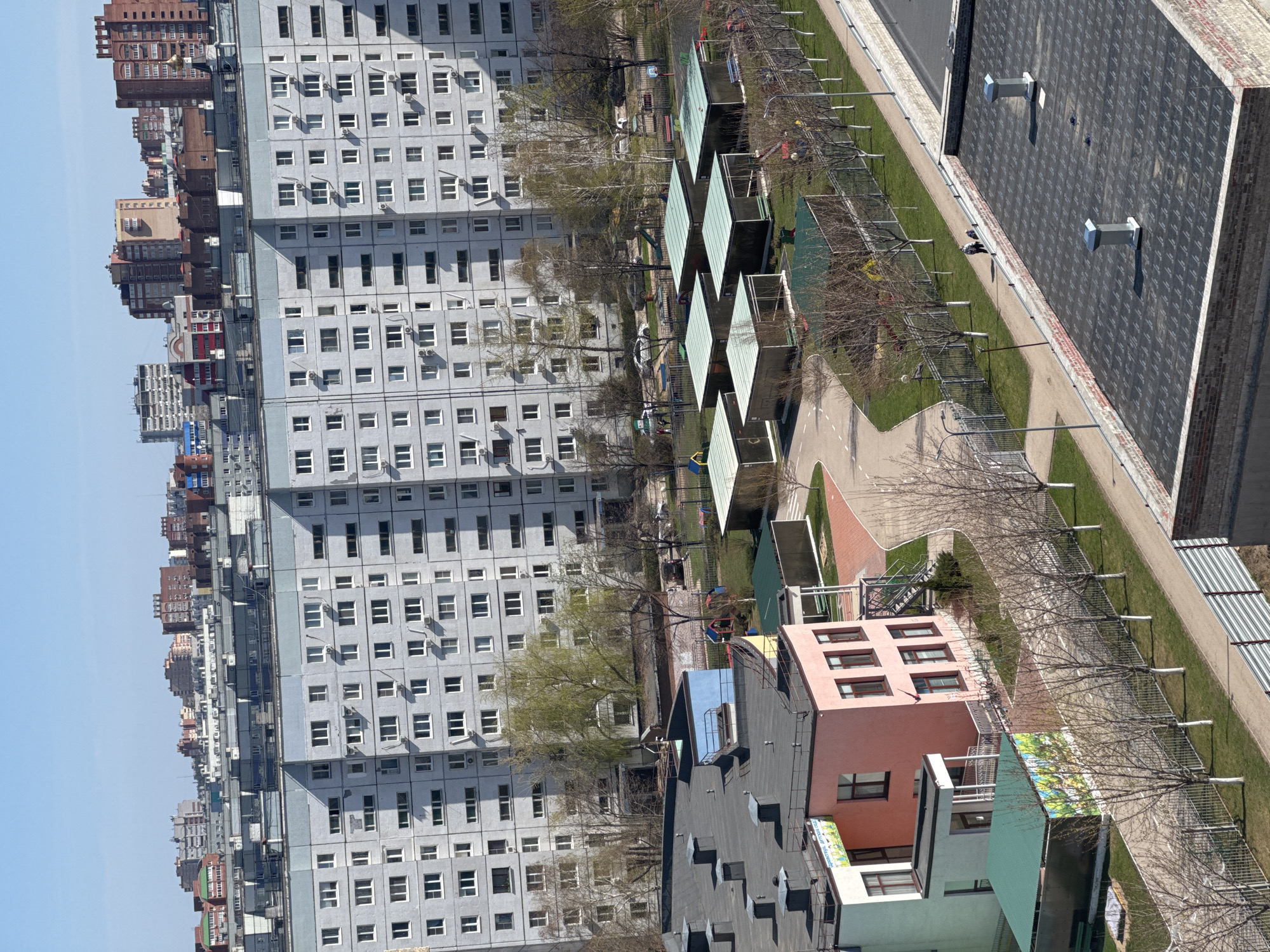 The image captures an expansive view of a residential urban landscape from an elevated perspective on a clear, sunny day. The dominant feature is a tall, multi-story gray apartment building with numerous windows and air conditioning units, stretching across the middle and left side of the frame. In the background, a dense skyline of other apartment buildings, varying in height and color (including brown, red, and lighter hues), recedes into the distance under a bright blue sky.
In the mid-ground, a green area with scattered trees, some bare and some showing early foliage, separates the large gray building from a paved path and an asphalt road or parking area. This green space contains a playground with equipment and several rectangular, modular structures with dark roofs, possibly kiosks or covered areas. Along the paved path, there are streetlights and metal fences.
In the foreground, two smaller, more modern-looking buildings are visible. One is a distinctively pink building with multiple windows, and adjacent to it is a mint-green building. The mint-green building features a colorful, abstract mural or sign on its side, but no discernible text is legible. The scene appears peaceful and quiet, with no people or active events visibly taking place, suggesting a calm daytime atmosphere, likely in early spring due to the condition of the trees. The overall impression is a typical residential district within a city.