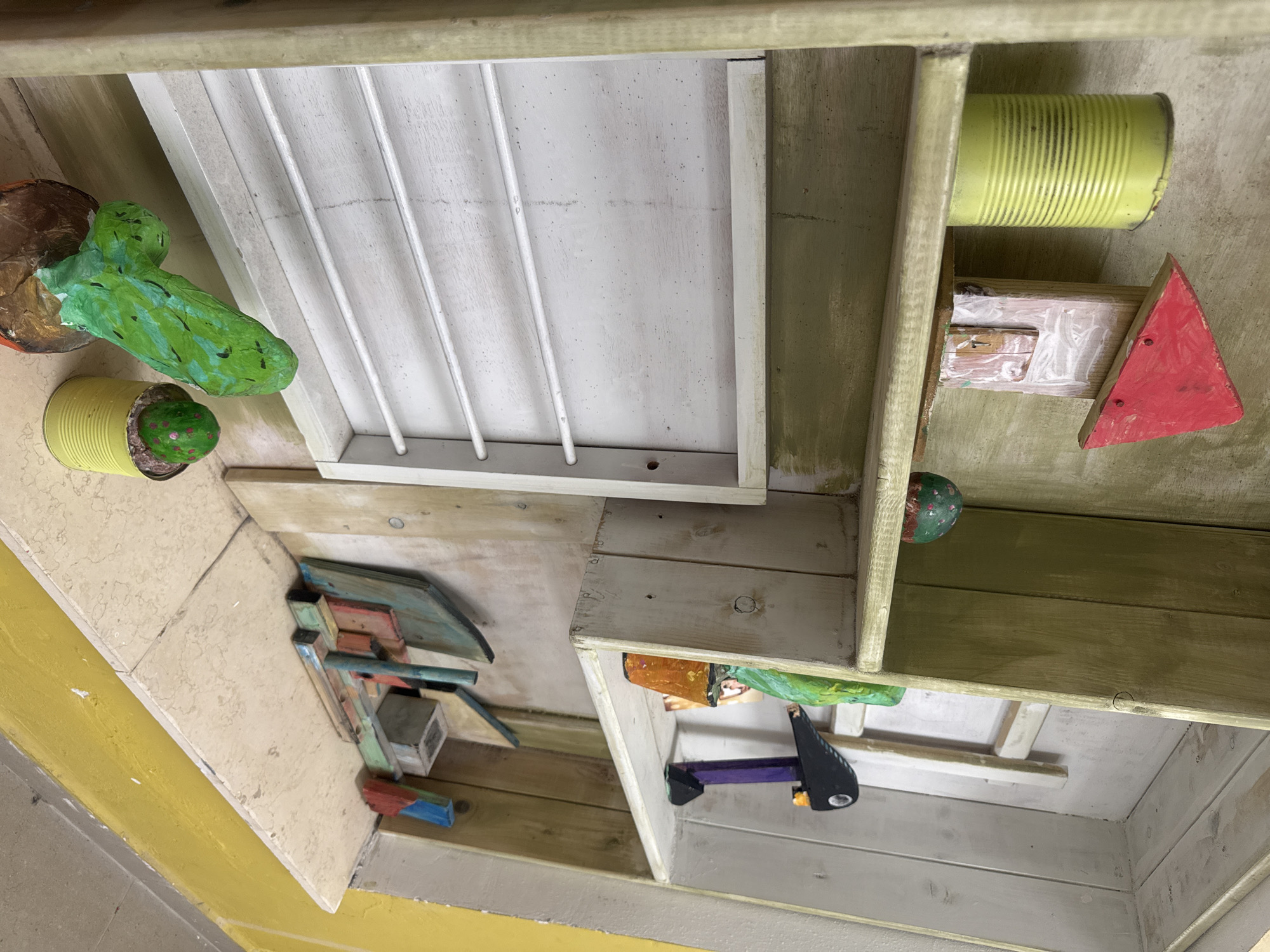 The image shows a multi-compartment wooden structure, possibly an art installation or a children's playset. It is located outdoors.
The compartments contain various objects including painted tin cans, geometric wooden shapes (a red triangle), a stapler, and what appears to be stylized cactus sculptures. The structure is painted in a variety of colors including yellow, green, red, and white. The wooden construction appears rustic with visible nail heads and paint imperfections. The image is rotated in an odd orientation. It could not be determined whether the object is in the city of Netanya, Israel.