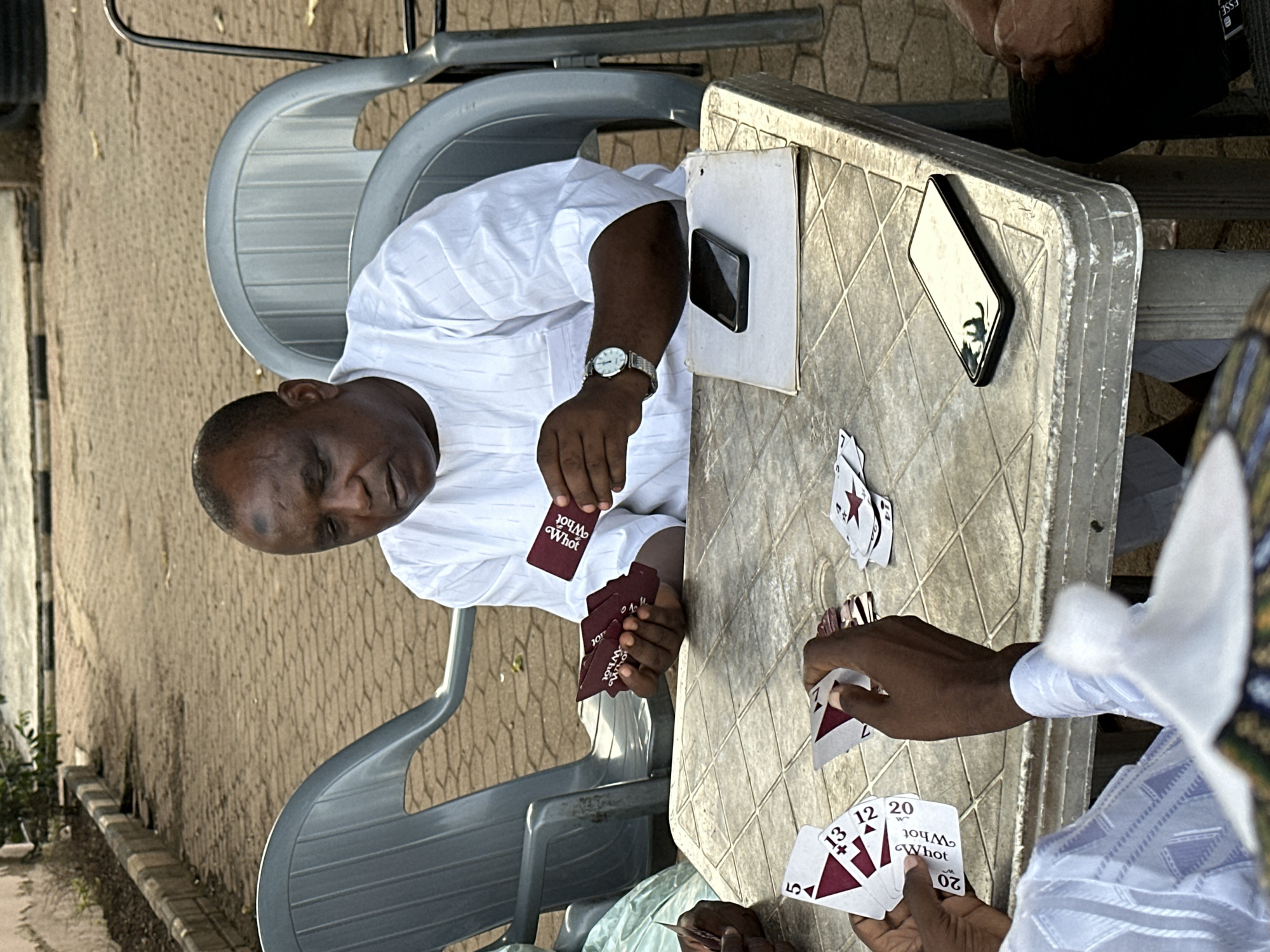 In Shaba, Nigeria, three individuals are engaged in a card game outdoors on a weathered plastic table. One man, wearing a white shirt, is seated to the left. Across from him, at least one other person holds a hand of cards. A third player is partially visible, wearing a patterned light blue and white shirt and a white cap. Playing cards are scattered on the table, along with two smartphones and a piece of paper. The setting appears to be on a paved surface with trees in the background. The lighting suggests it is daytime.