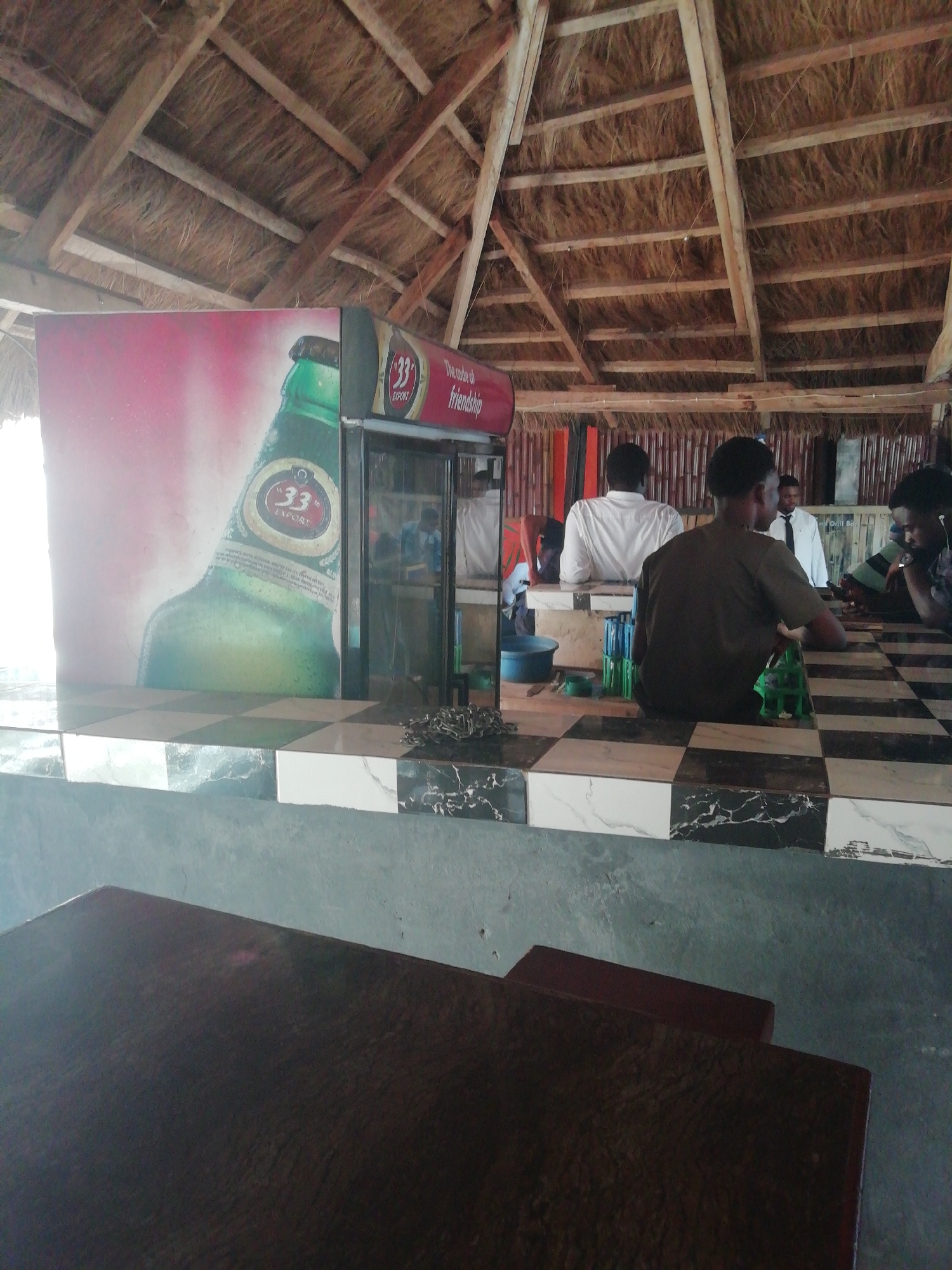 The image shows the interior of an open-air bar in Enugu, Nigeria. The building has a thatched roof. A refrigerator with an advertisement for "33" Export beer is visible, partially blocking the view. Several men are seated at a checkered-tile countertop. Some appear to be engaged in conversation. A dark wooden table dominates the foreground. A set of keys and several green plastic crates are on the countertop.