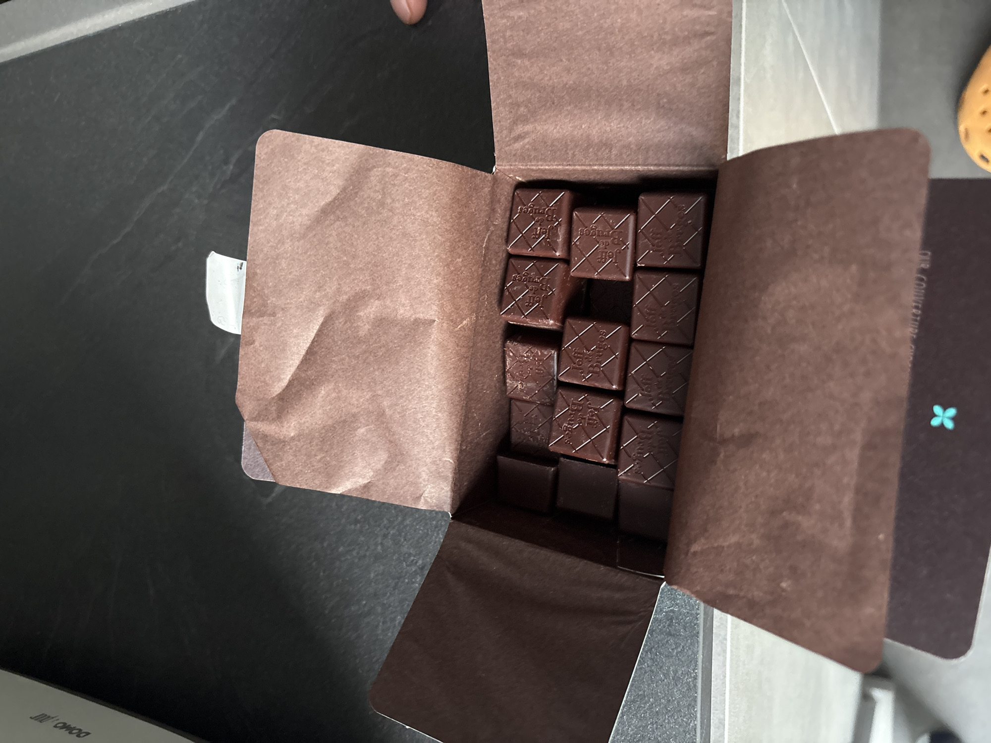 A box of chocolates sits open on a countertop of dark gray. The box is brown and contains multiple square chocolates with a decorative pattern on each. The box is partially open, revealing the chocolates inside. A portion of the box has a small teal-colored symbol on it. To the left of the box, the surface is a dark countertop. The scene appears to be indoors, possibly in a kitchen. The location is Unknown, Unknown.