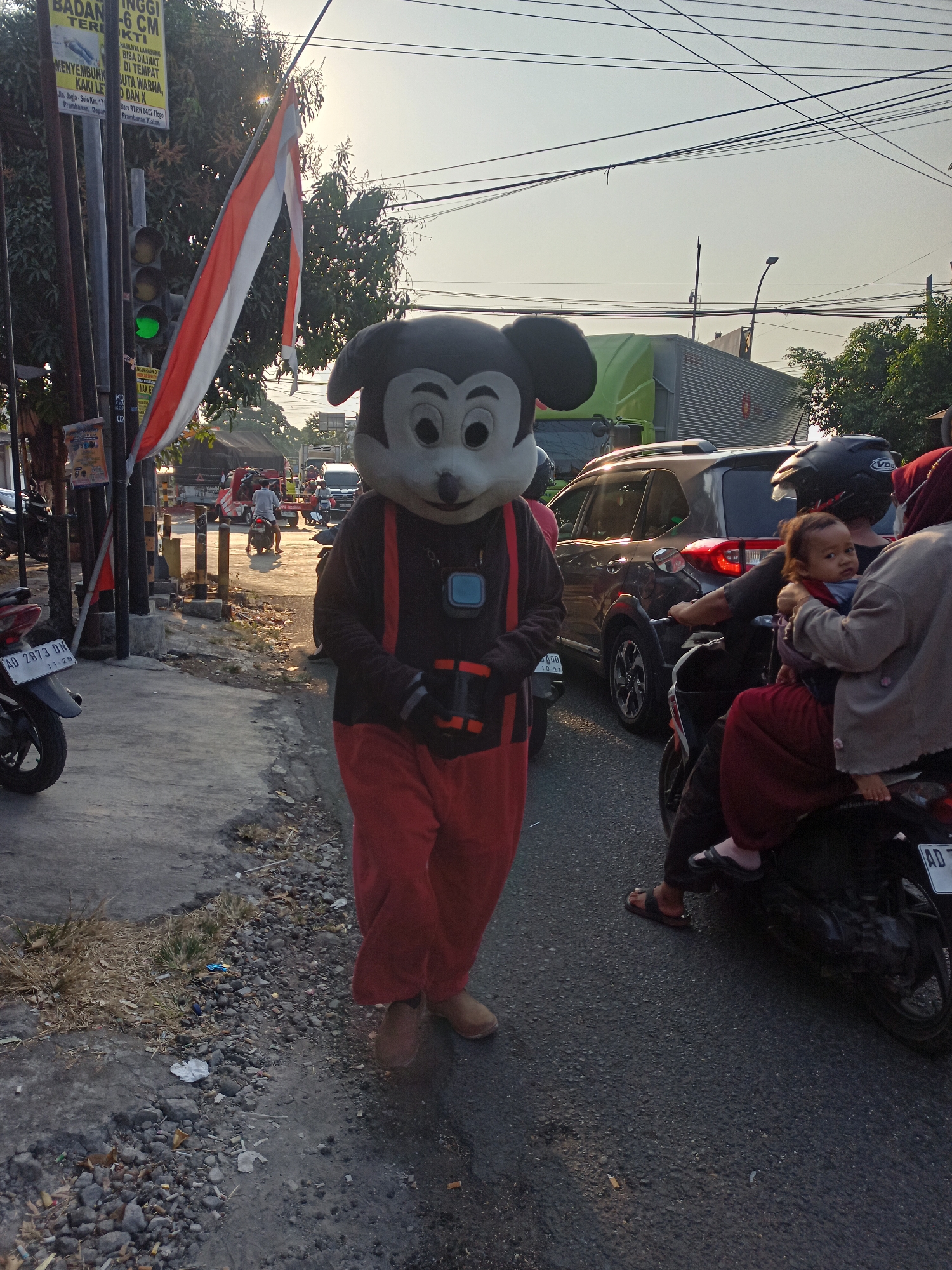 A Mickey Mouse mascot costume wearer, with red pants and black suspenders, walks along the side of a street in Gemolong, Indonesia. The mascot's face is large and white with black ears. A child is riding on the back of a motorcycle with an adult, who is wearing a red hijab. The street is paved and has traffic. There are trees and power lines visible in the background. A traffic light shows a green signal. The sun is low in the sky, casting long shadows. The scene appears to be taking place during the daytime.