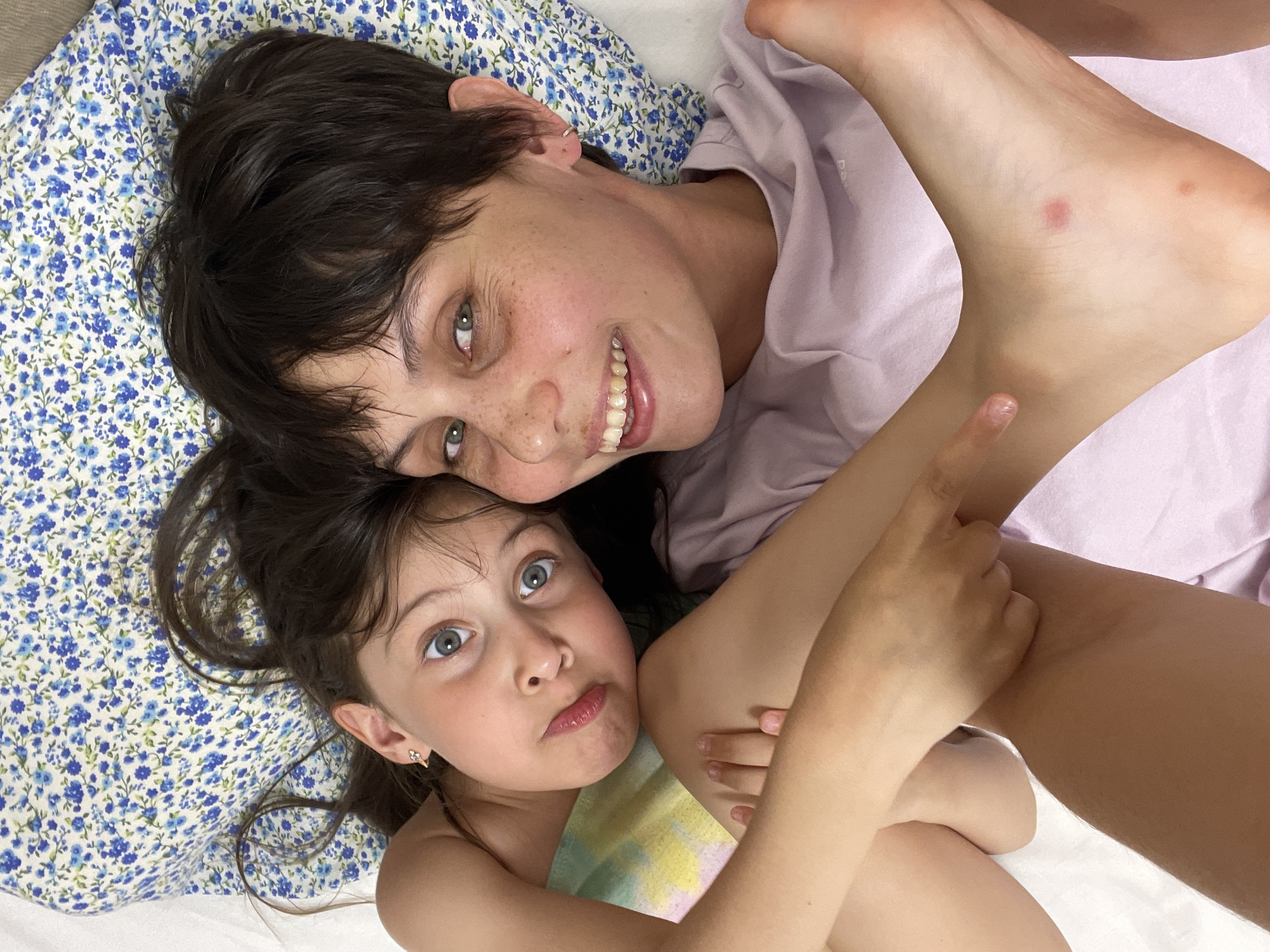 Two individuals, presumably a mother and daughter, are lying down. The mother is positioned on a pillow with a blue floral pattern. She has short, dark hair, fair skin with freckles, and is smiling. She is wearing a light purple t-shirt. The daughter is lying next to her with long, dark hair and fair skin. She has an uncertain expression. The daughter is wearing an unseen garment with a tie-dye pattern. Limbs are intertwined between the two individuals. The location is unspecified, and there's no indication that it is in Izhevsk, Russia.